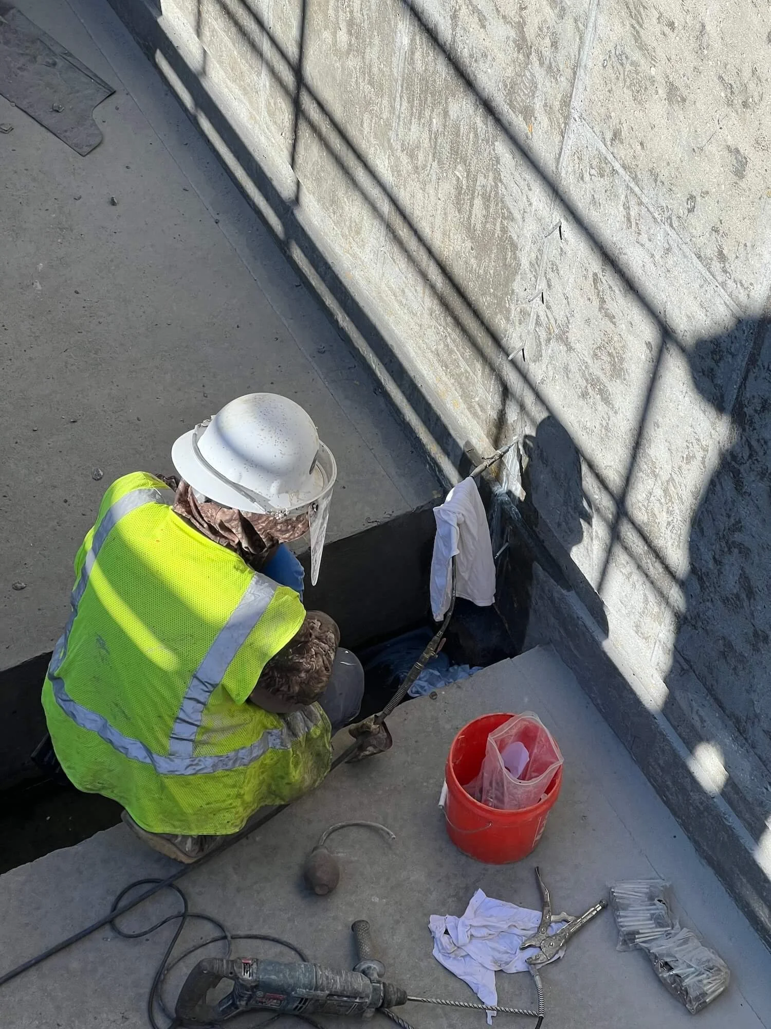 Worker in a yellow safety vest and white helmet working on a construction site near a concrete wall, with tools and equipment on the ground.