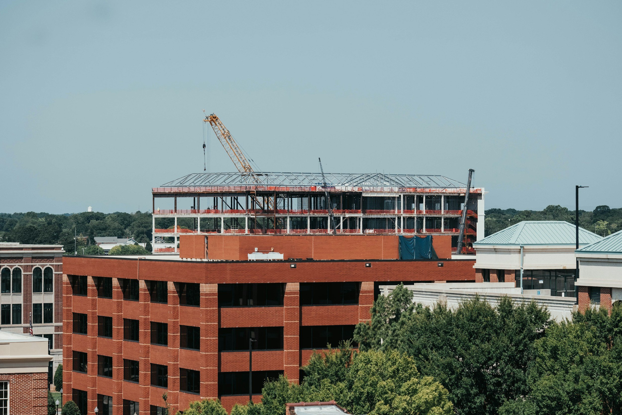A multi-story brick building under construction with a crane on top, surrounded by trees and other buildings, with a clear blue sky in the background.