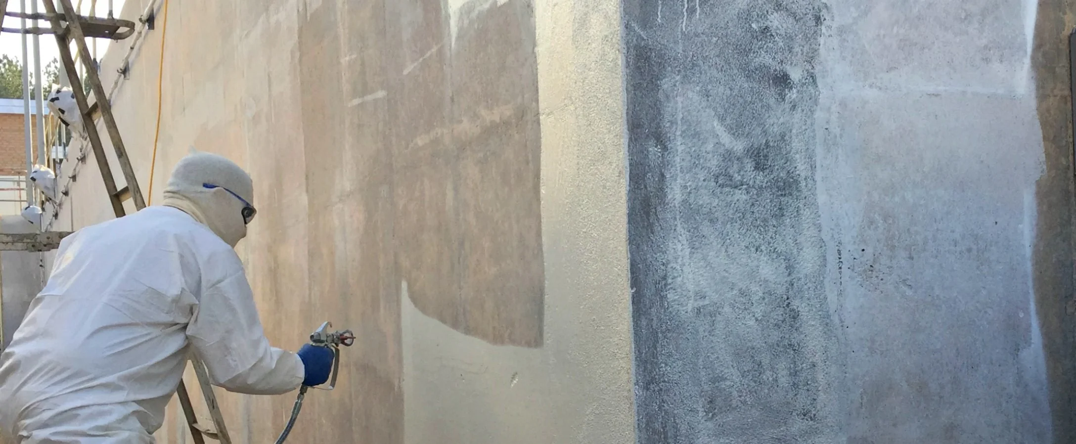 A person wearing white protective clothing, gloves, and a face mask spray painting a wall.