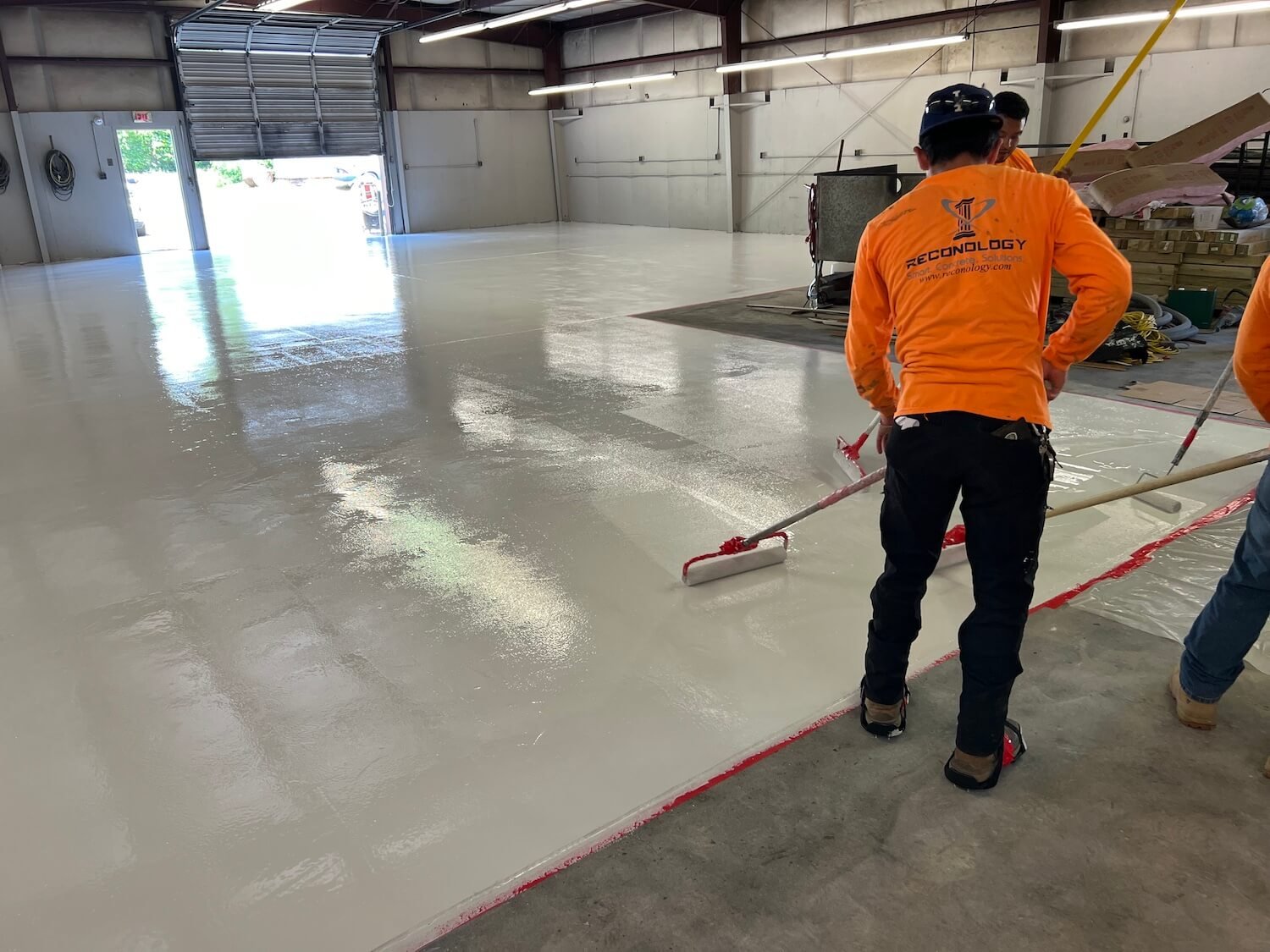reconology workers in orange shirts are applying a fresh layer of epoxy coating on the concrete floor inside a large warehouse or industrial building.