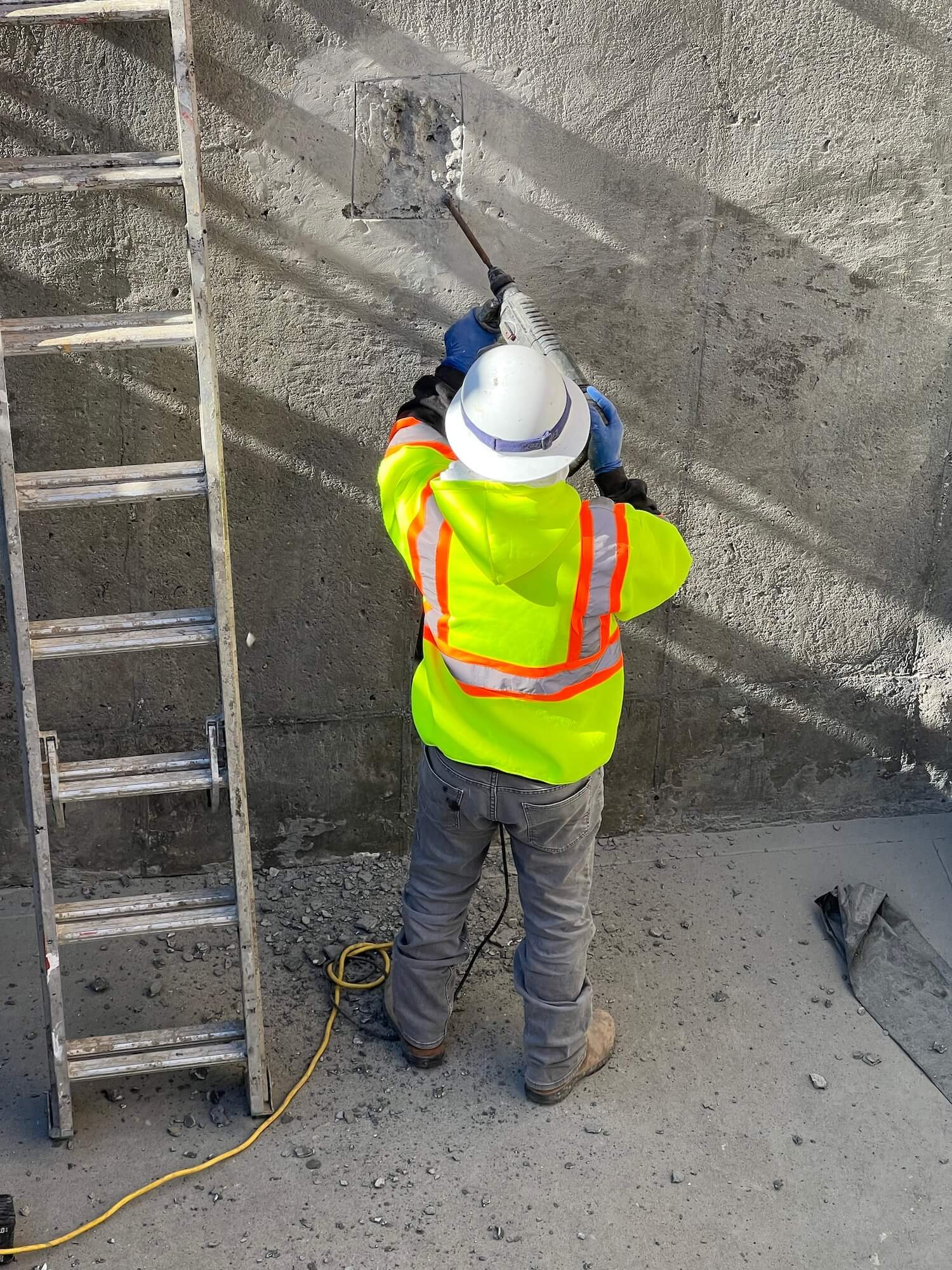 A construction worker wearing a white hard hat, a reflective safety vest, and gray pants is using a power tool on a concrete wall.