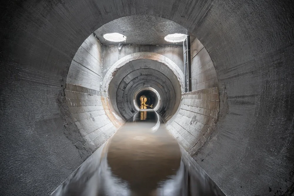 Underground tunnel with a flowing water drain and circular openings overhead.