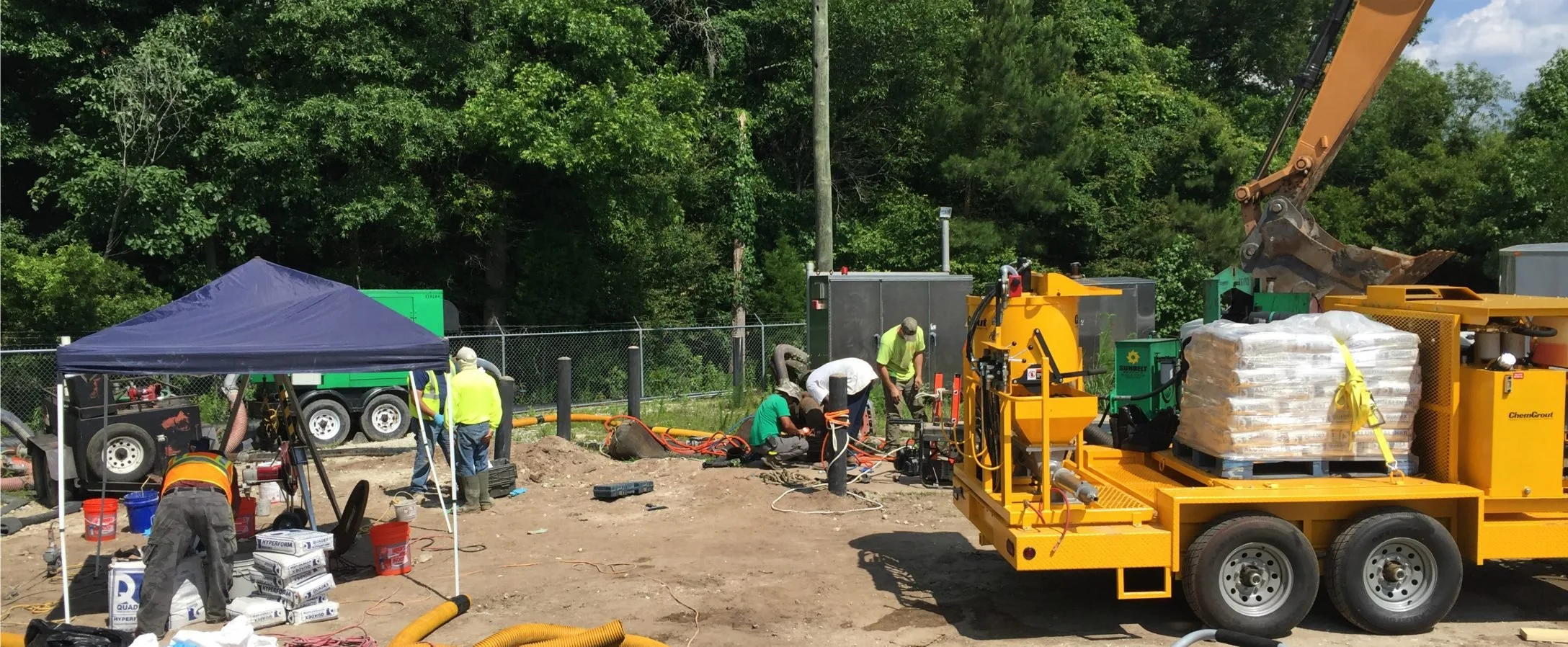 Workers in safety vests and helmets working on a construction site with machinery, equipment, and a cyan tent, near a chain-link fence and a wooded area.