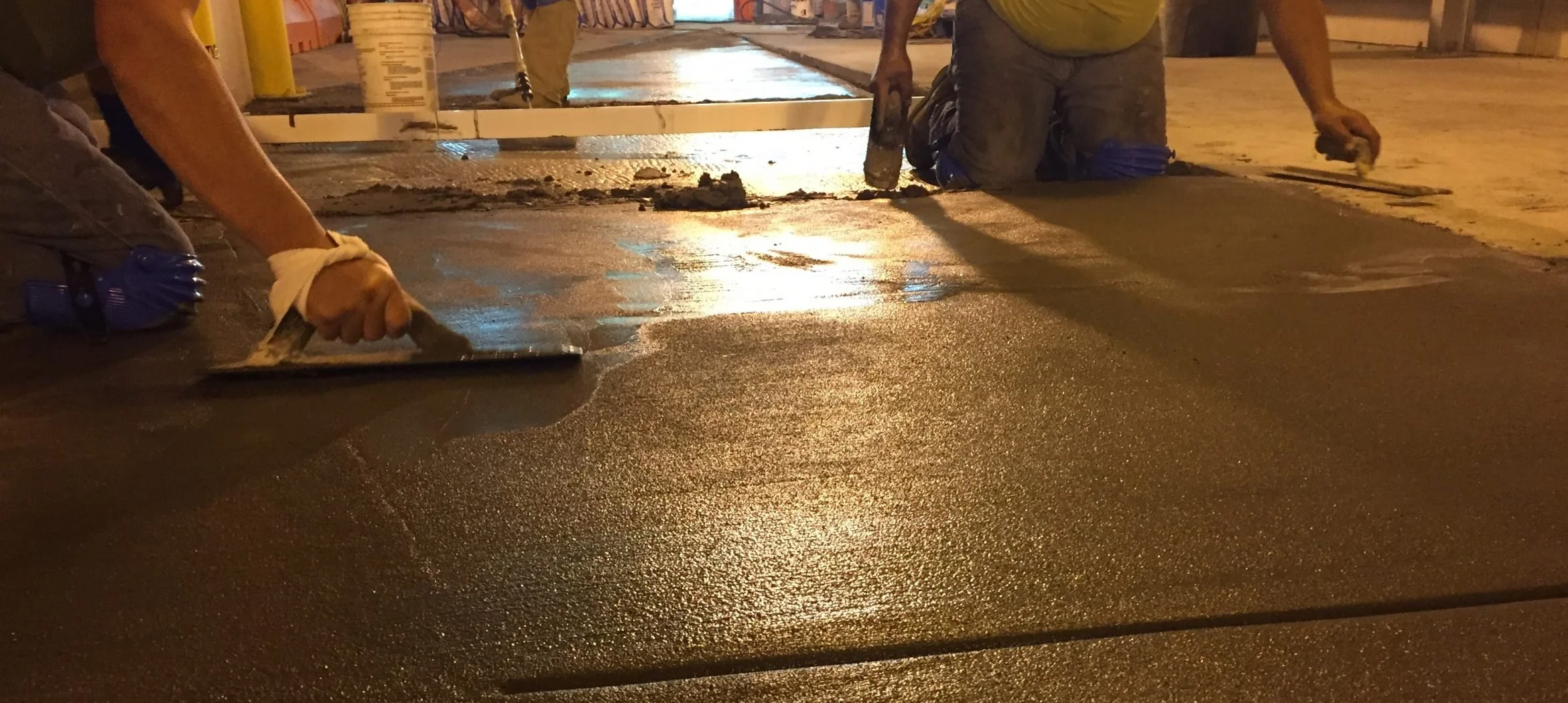 Workers applying wet concrete to a sidewalk at night, kneeling and smoothing the surface with trowels.