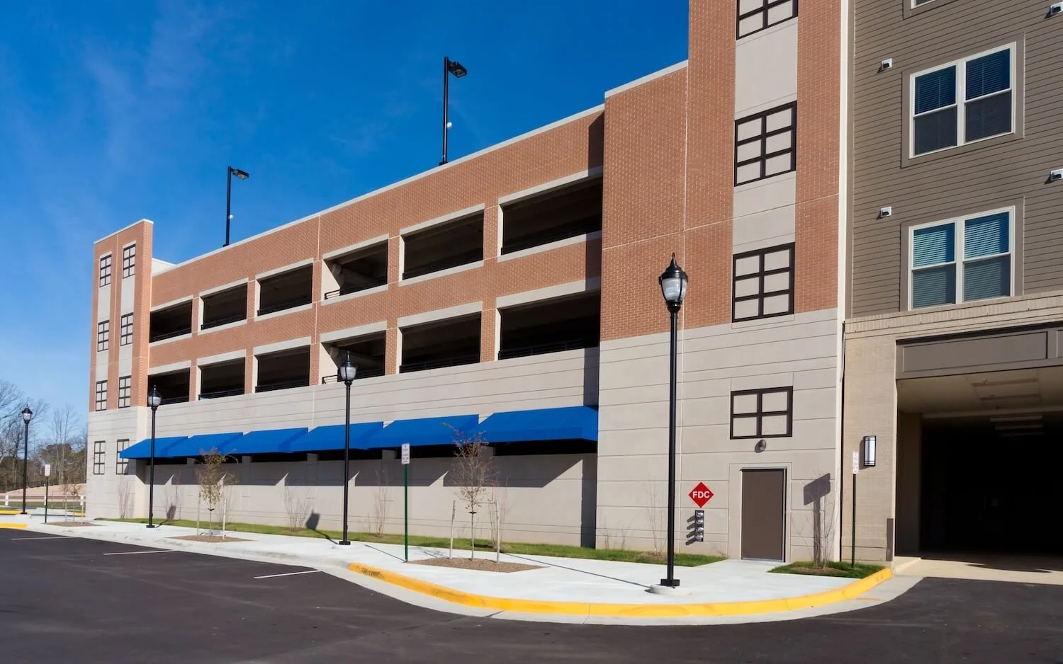 Exterior view of a multi-story parking garage with brick and beige walls, blue awnings, street lamps, small trees, and a clear blue sky.