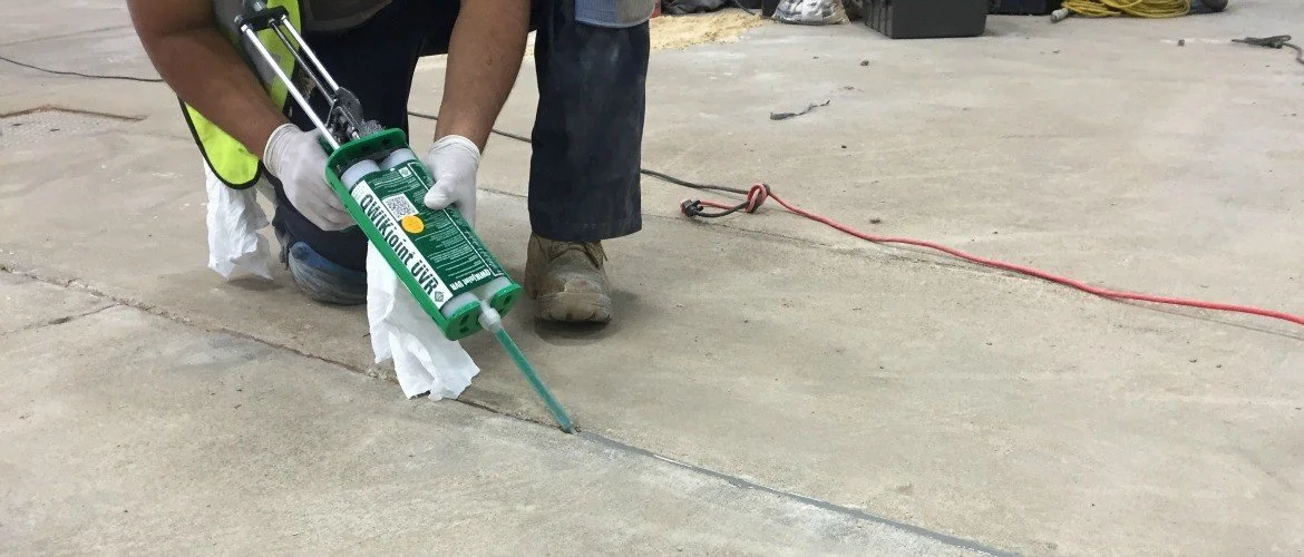 A worker kneeling on the concrete floor, applying caulk along a seam with a caulk gun. The worker is wearing gloves, a safety vest, and work boots. Various tools and equipment are visible in the background.
