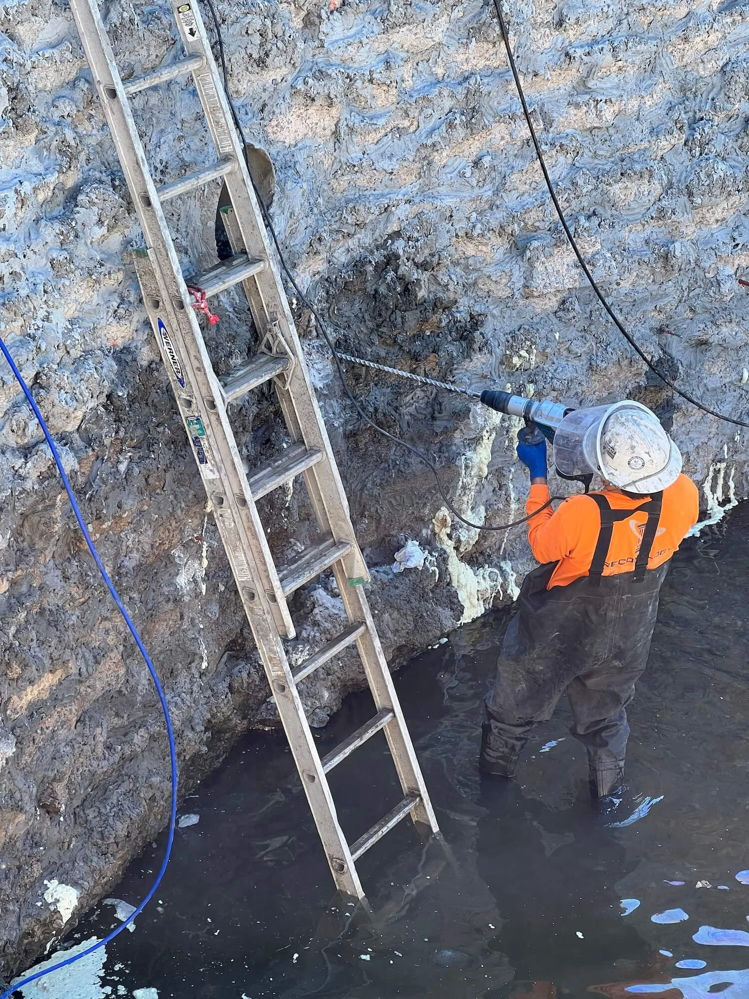 A worker in an orange jacket, gloves, and helmet standing in water next to a rocky wall, holding a power tool, with a tall silver ladder leaning against the rocks nearby.