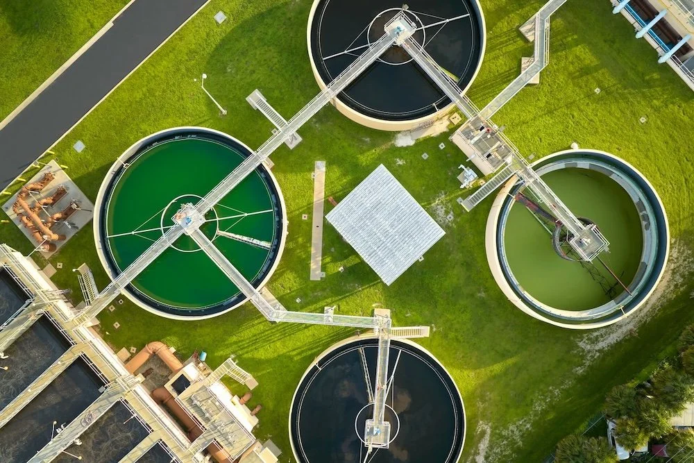 Aerial view of a water treatment facility with four circular sedimentation tanks connected by walkways and pipelines, situated on a grassy area with some infrastructure nearby.