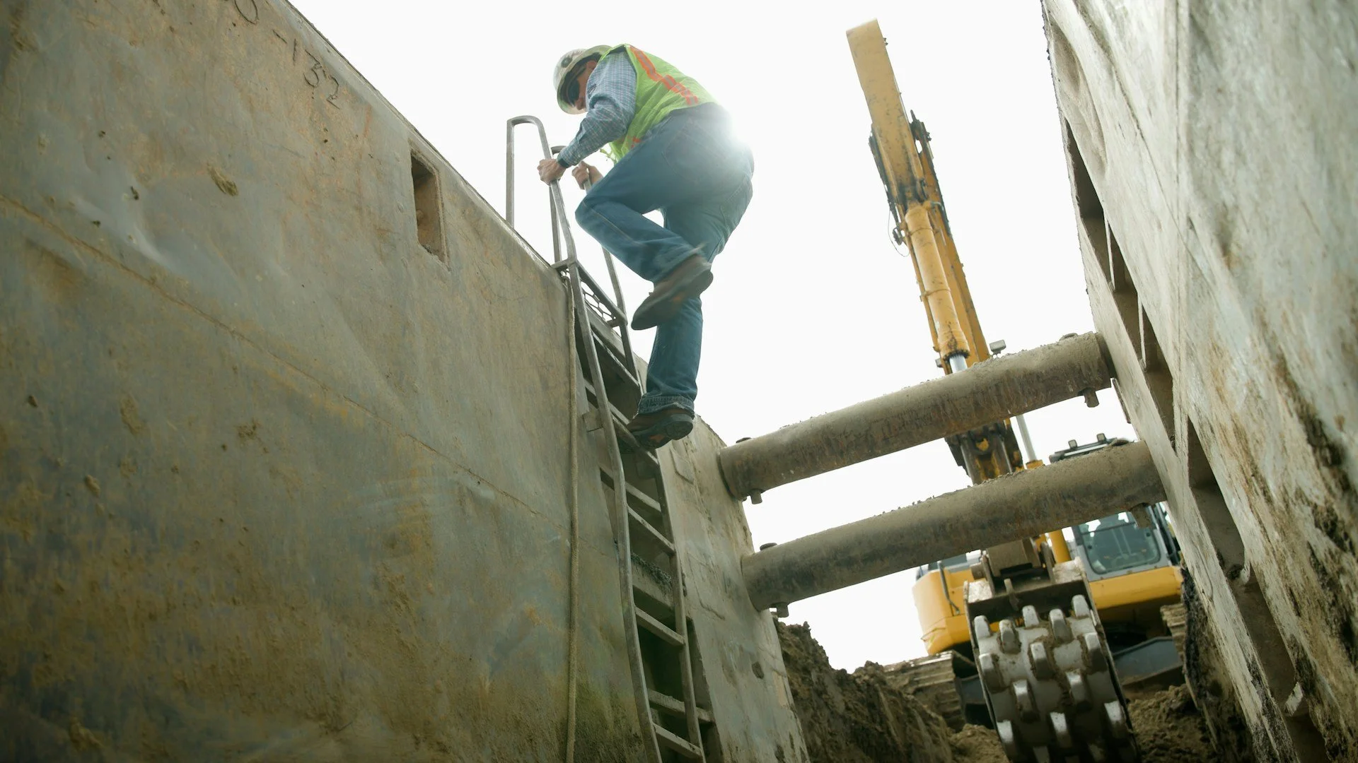 Construction worker in safety gear climbing a ladder into an excavation site with heavy machinery in the background.