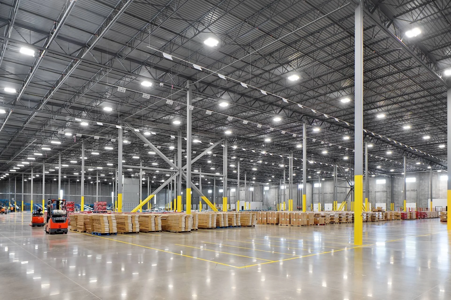 Interior of a large warehouse with high ceiling, bright overhead lighting, and yellow support poles. Floor is spacious and organized with pallets of boxes and orange forklifts.