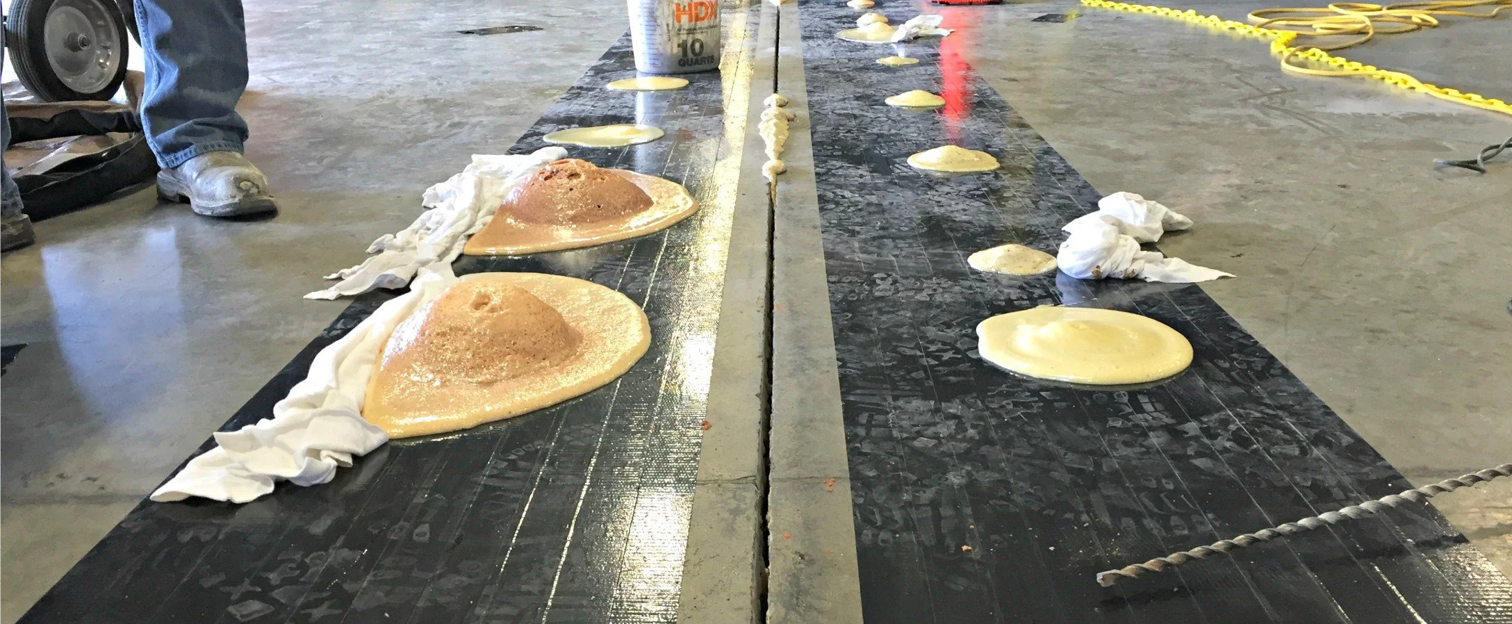 A concrete floor with two black conveyor belts side by side, each with large, bubbly, light-colored foam blobs. There are some white cloths, a can of spray foam, and person’s legs wearing jeans and work boots visible in the background.