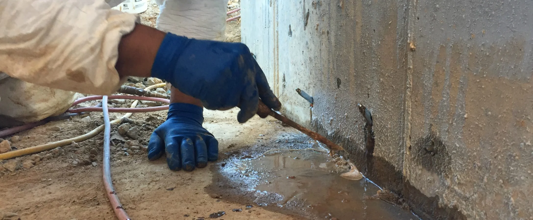 A person wearing blue gloves and work clothes is using a tool to repair a pipe underneath a concrete surface. There are several hoses and pipes around, indicating a construction or plumbing repair site.