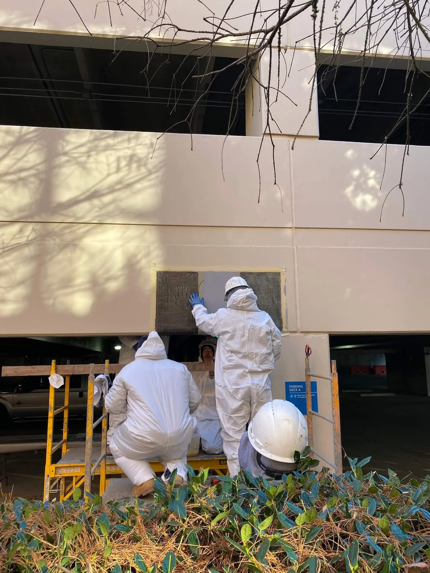 People dressed in white protective suits, helmets, and gloves working on or inspecting a area under an overpass, with some greenery in the foreground and a parking garage in the background.