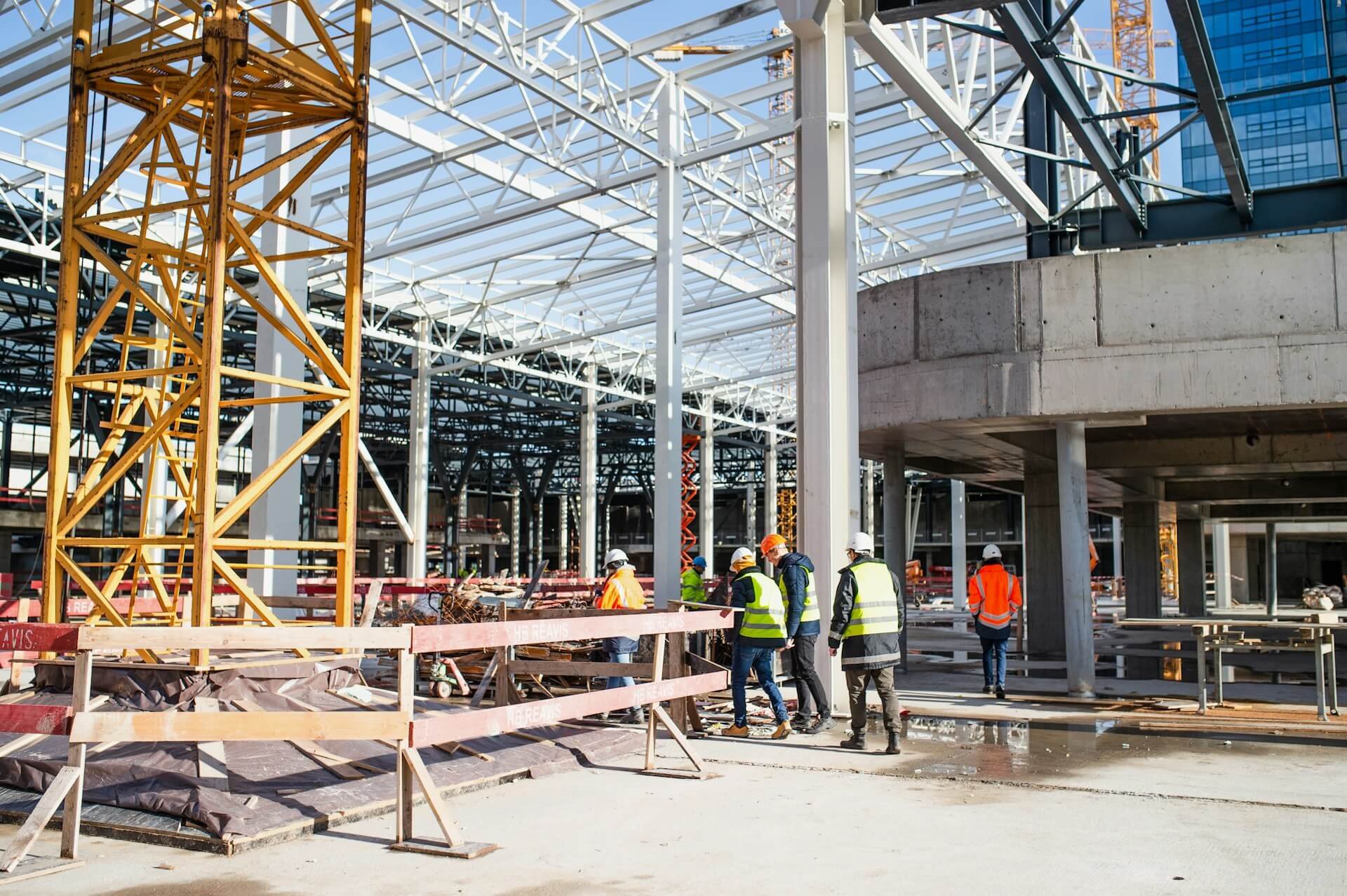 Construction site with workers wearing safety gear, metal framework, a large yellow tower crane, and an unfinished building structure.