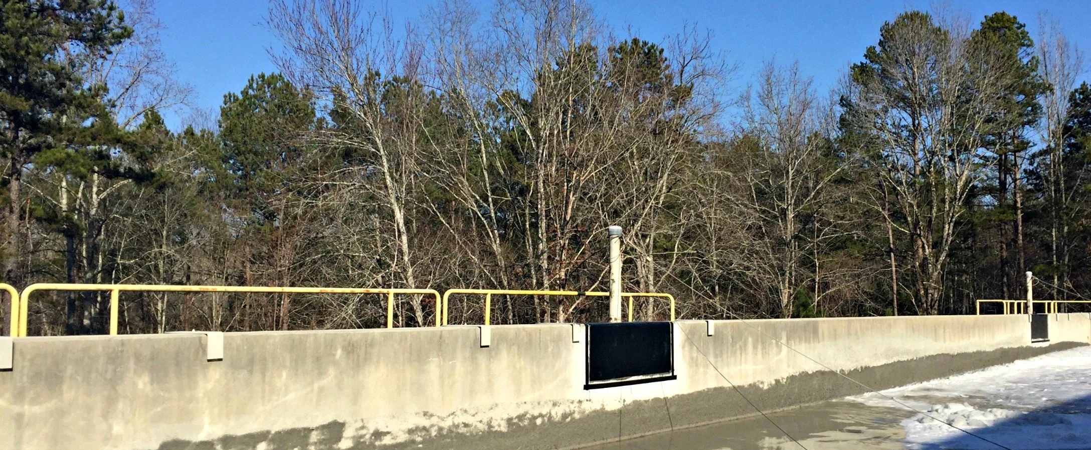 A concrete structure with yellow railings and a black gate, set against a backdrop of leafless trees and evergreens under a clear blue sky, with snow on the ground.