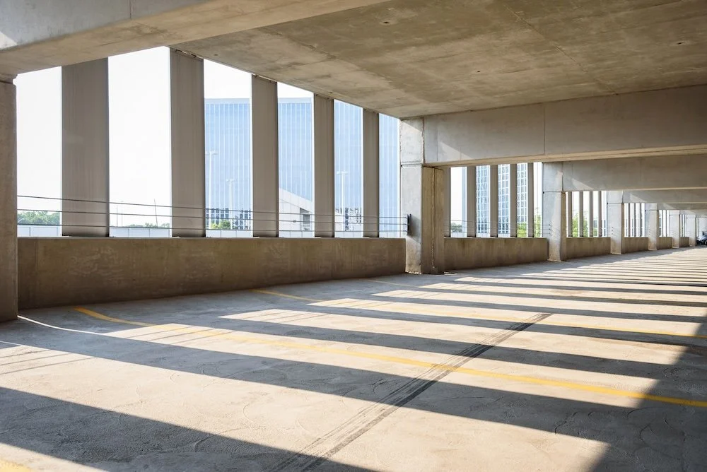 An empty parking garage with sunlight casting shadows, concrete floors and pillars, and large windows with city buildings outside.