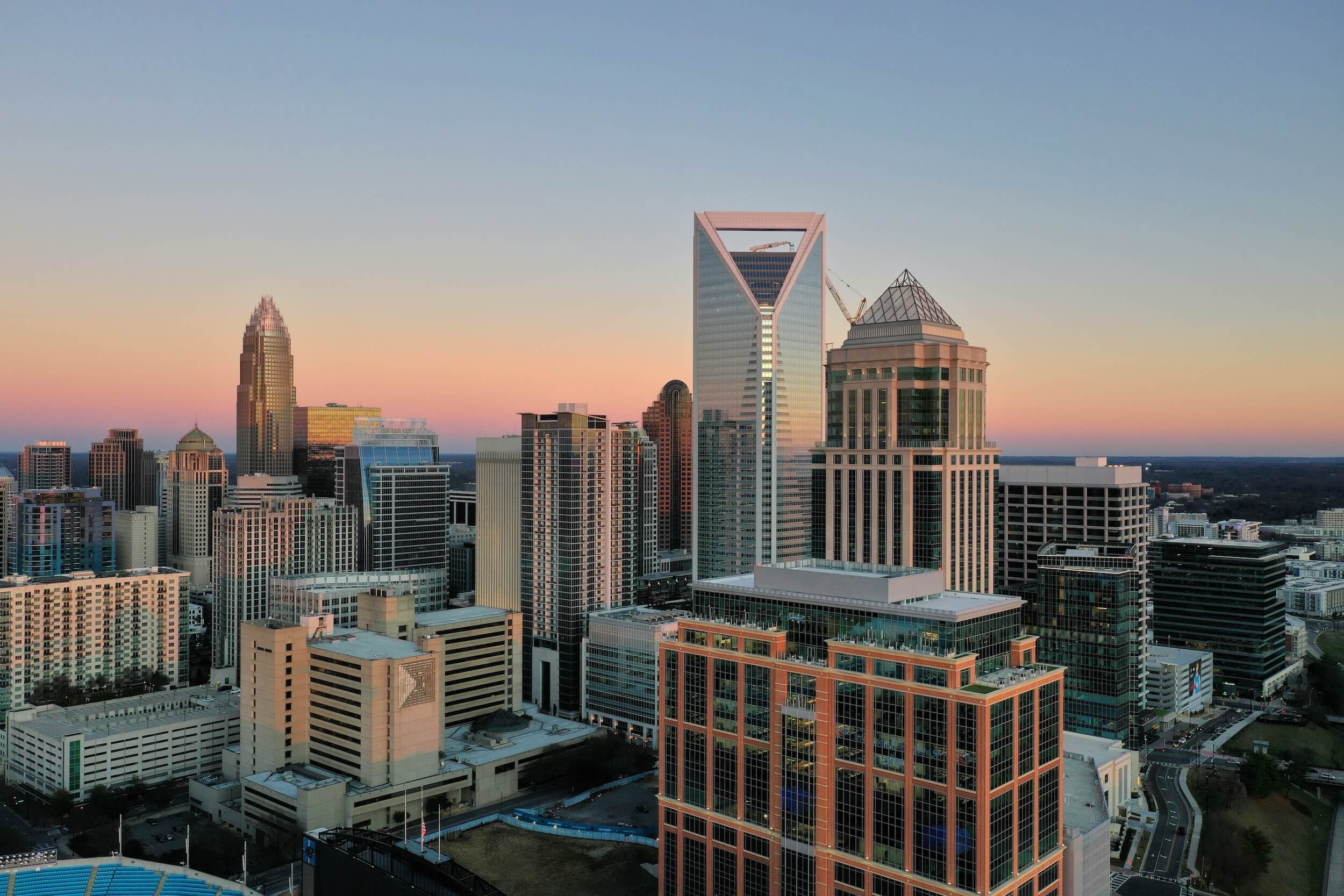 A city skyline during sunset featuring tall modern skyscrapers, including one with a distinctive triangle-shaped top, and a background of a clear sky fading from pale orange to blue.