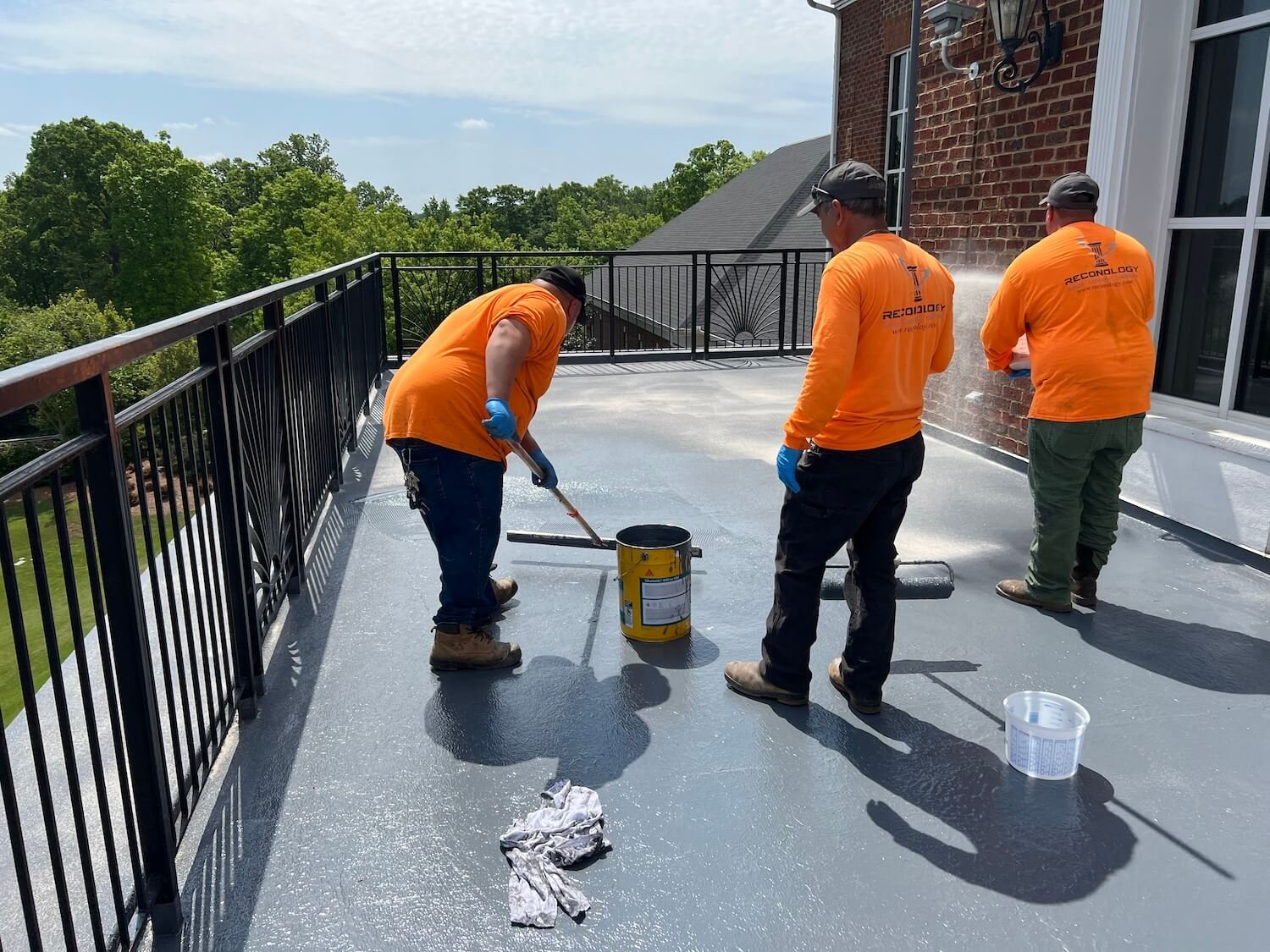 Four reconology workers in orange shirts applying waterproof coating to a rooftop patio, with a black metal railing, green trees in the background, and a brick building to the right.