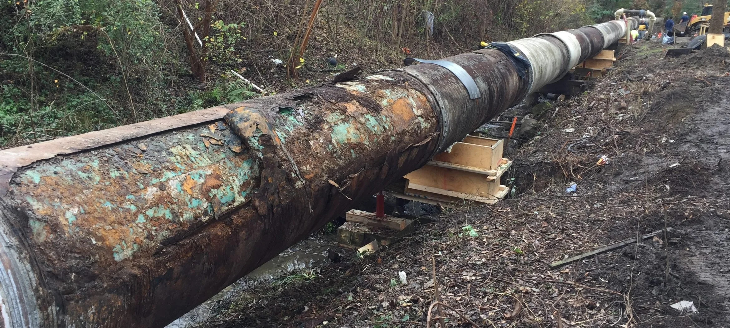 A large, rusty pipe in a wooded area undergoing maintenance or repair, supported by wooden blocks and surrounded by dirt and debris.