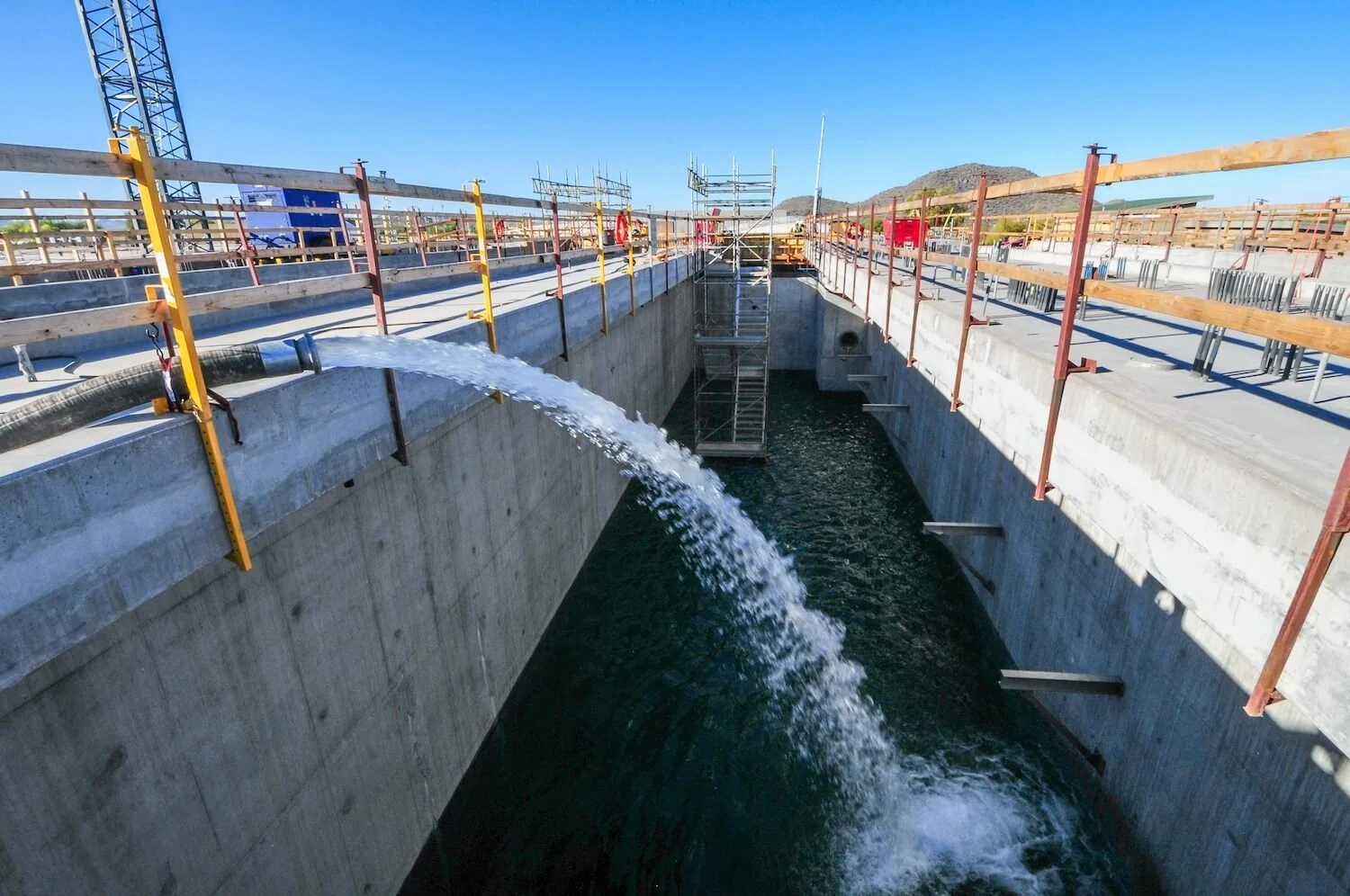 Construction site with a concrete canal, water flowing out of a pipe, metal scaffolding, safety barriers, and clear blue sky.