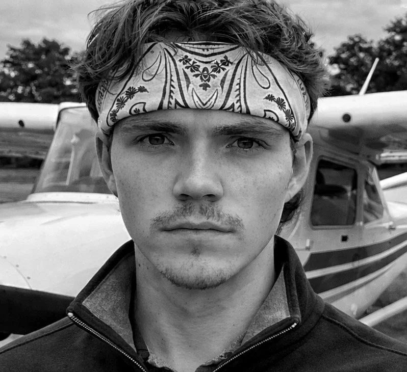 A young man with a bandana on his forehead standing in front of a small airplane.