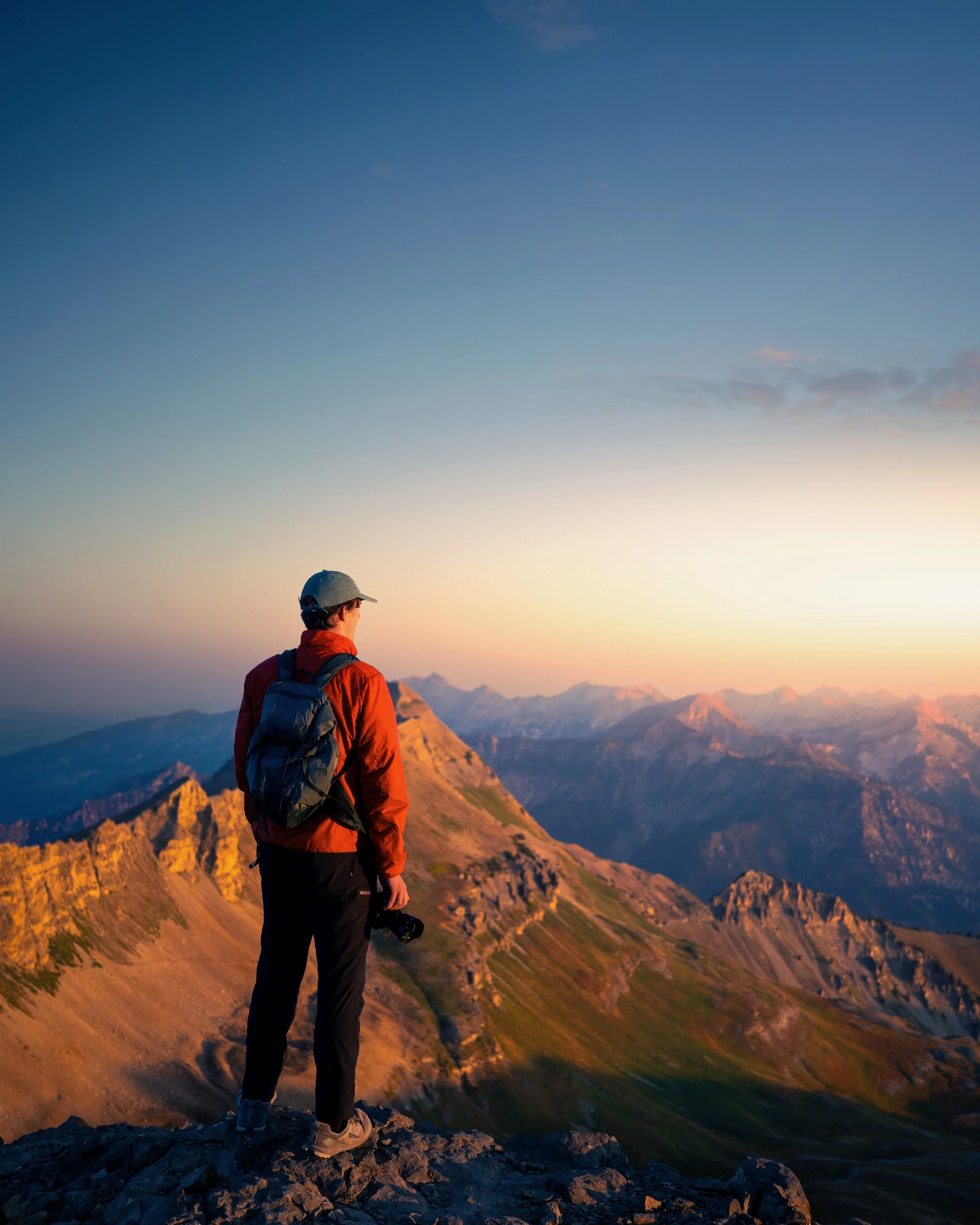 A person dressed in outdoor gear, including a baseball cap and a backpack, standing on rocky terrain overlooking a mountain range during sunset or sunrise, with a clear sky and distant peaks.