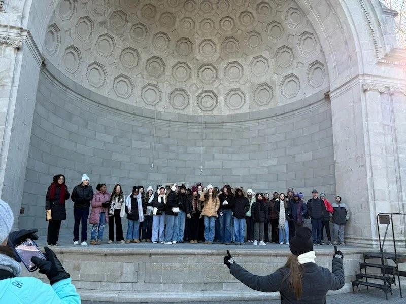 A group of people standing on a stage in front of a large stone wall with decorative patterns, possibly at a historical site or monument.