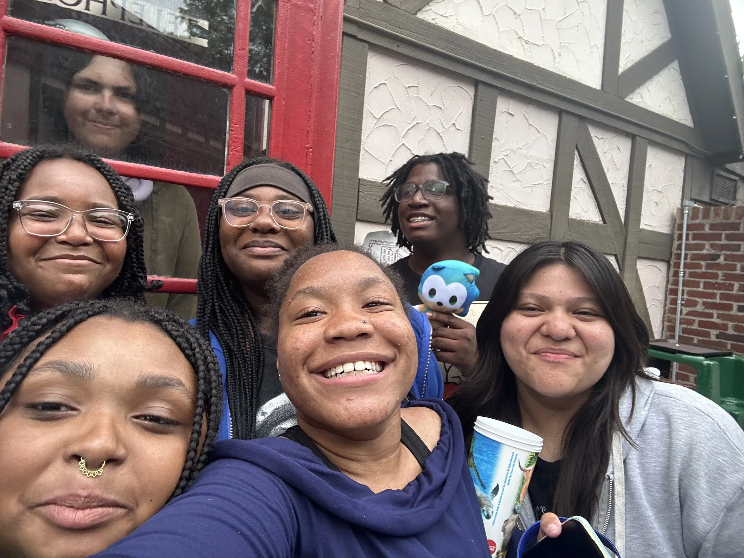 A group of diverse smiling people taking a selfie outdoors in front of a building with a red window frame and brick wall. One person is holding a colorful plush toy.