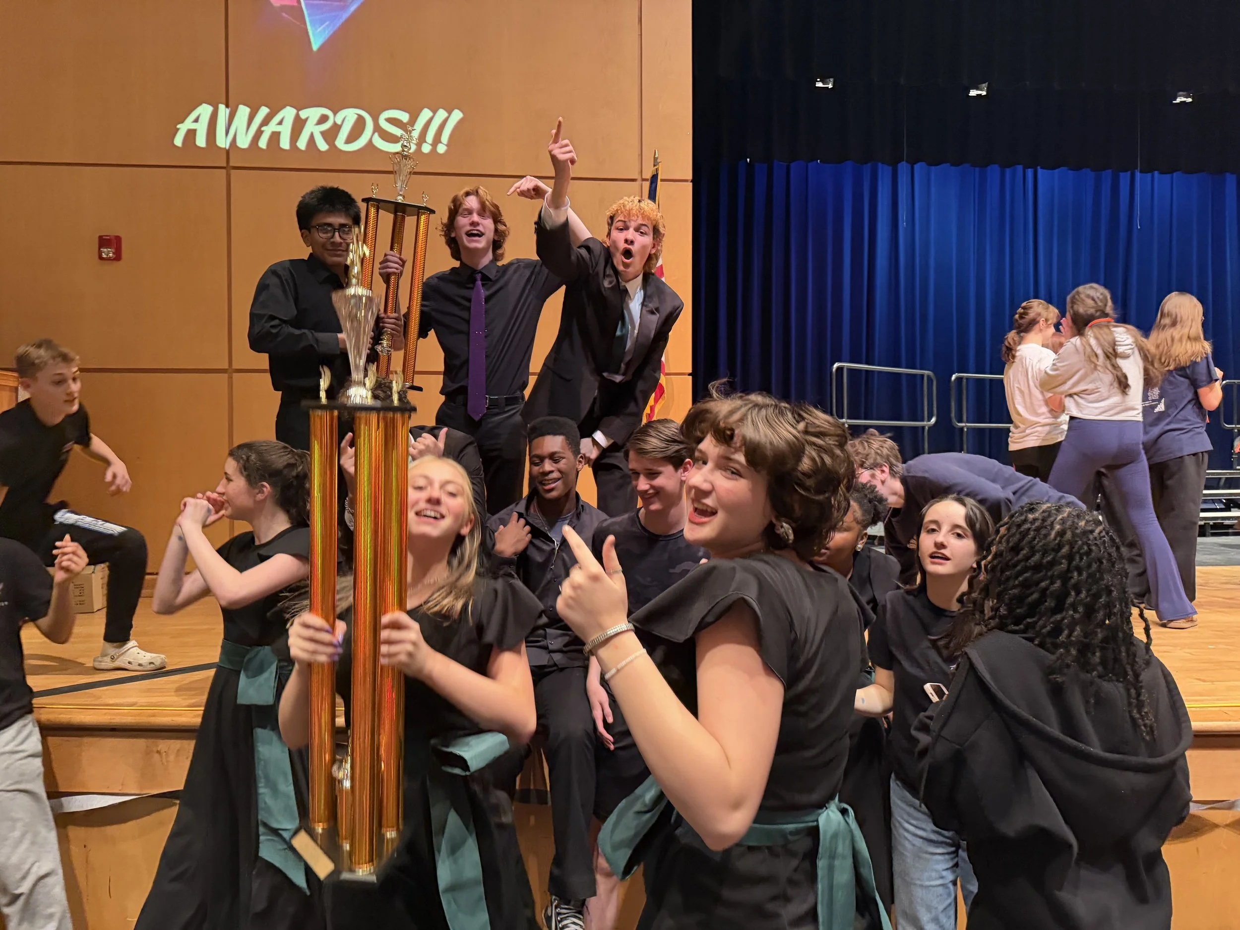 A group of students on stage celebrating at an awards ceremony, with three students holding large trophies in the center. The backdrop features a wooden wall and a blue curtain, with a large projection on the wall that reads 'AWARDS!!'.
