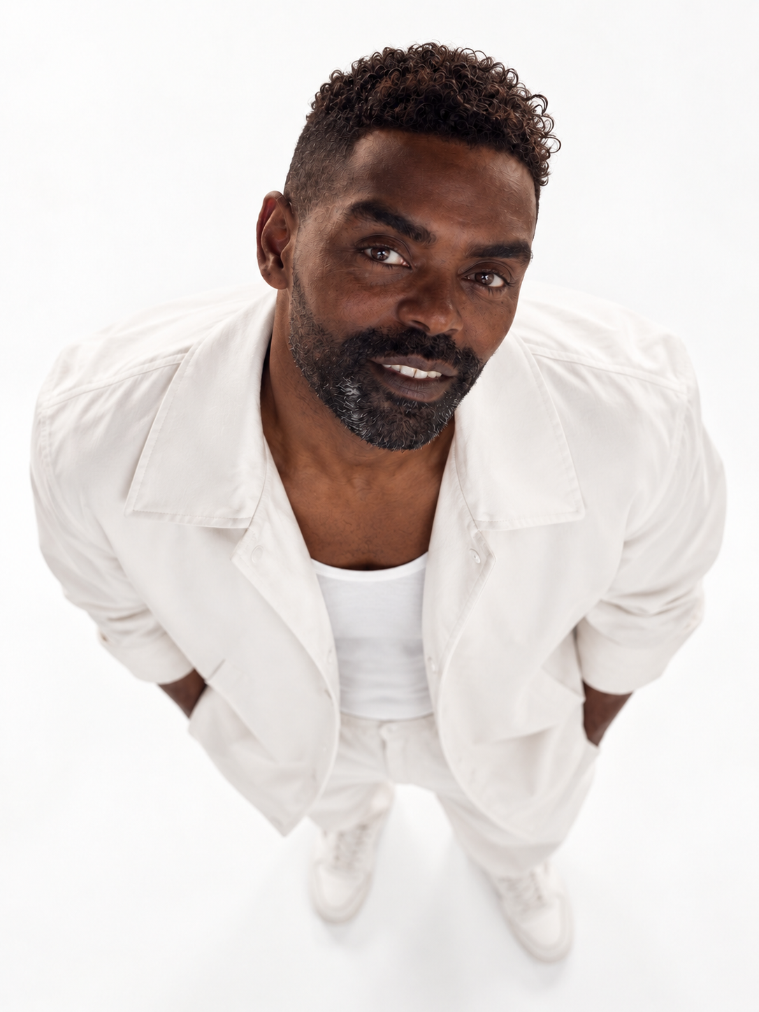 A man with curly hair and a beard, dressed in white, looking up toward the camera with his hands behind his back.
