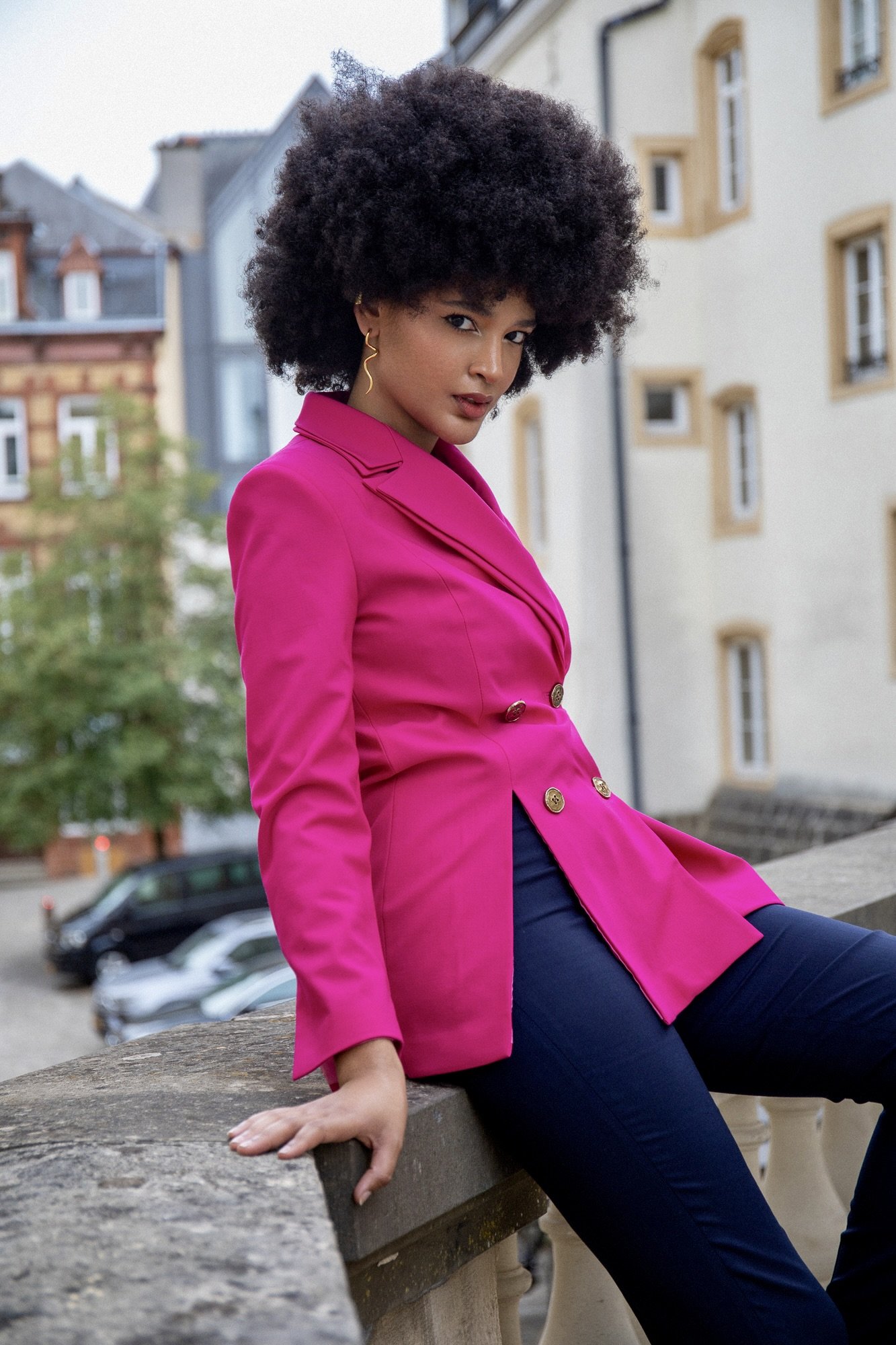 Urban fashion editorial photographed by Henri Da Cruz in Luxembourg City, featuring model Ana wearing a vibrant fuchsia blazer by Francini_K with dark tailored trousers. A relaxed seated pose on a stone balustrade highlights the structured silhouette