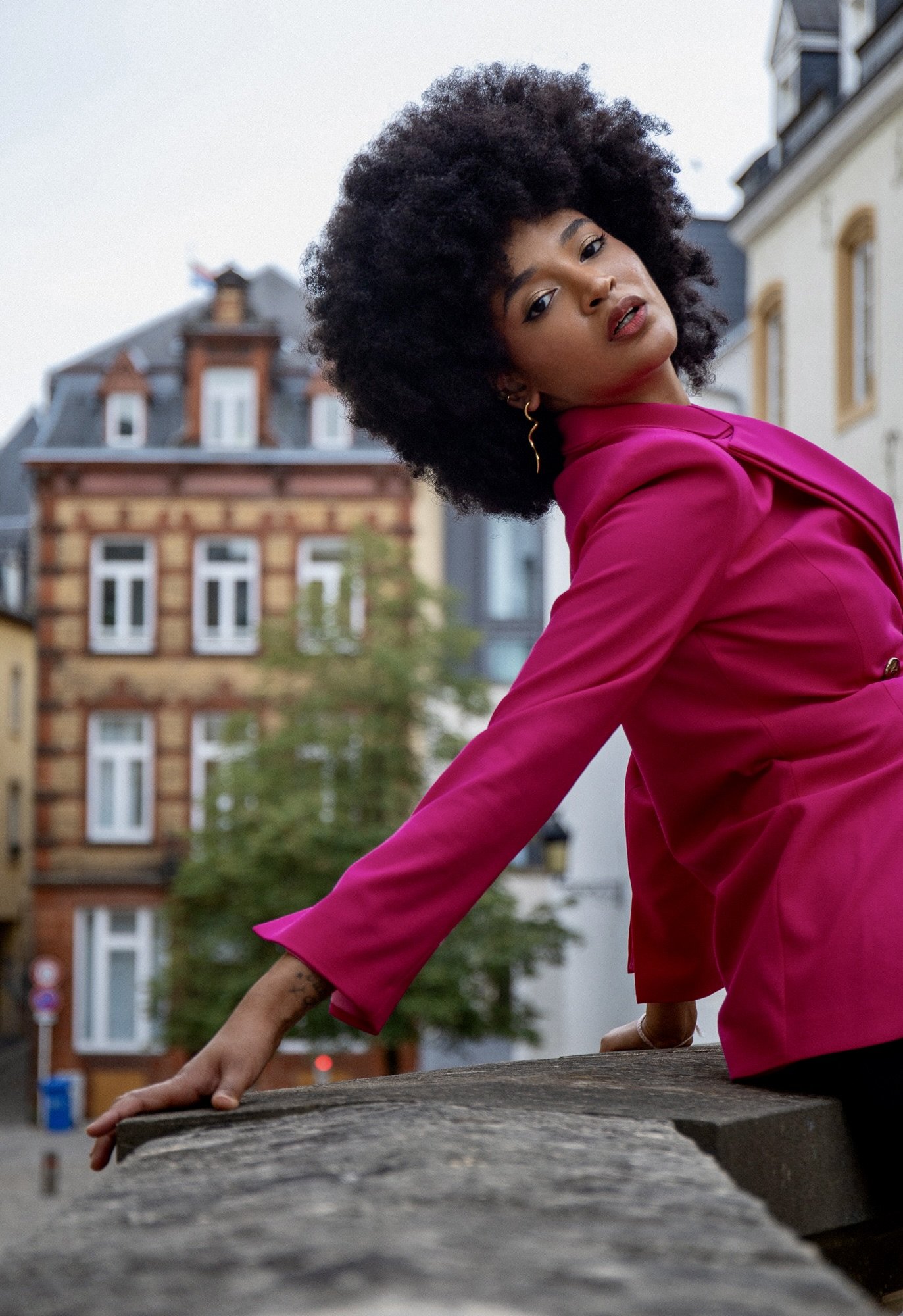 Editorial fashion photography by Henri Da Cruz in Luxembourg City, featuring model Ana in a vibrant fuchsia blazer by Francini_K. A dynamic leaning pose against a stone balustrade combines bold contemporary tailoring, expressive movement and elegant 