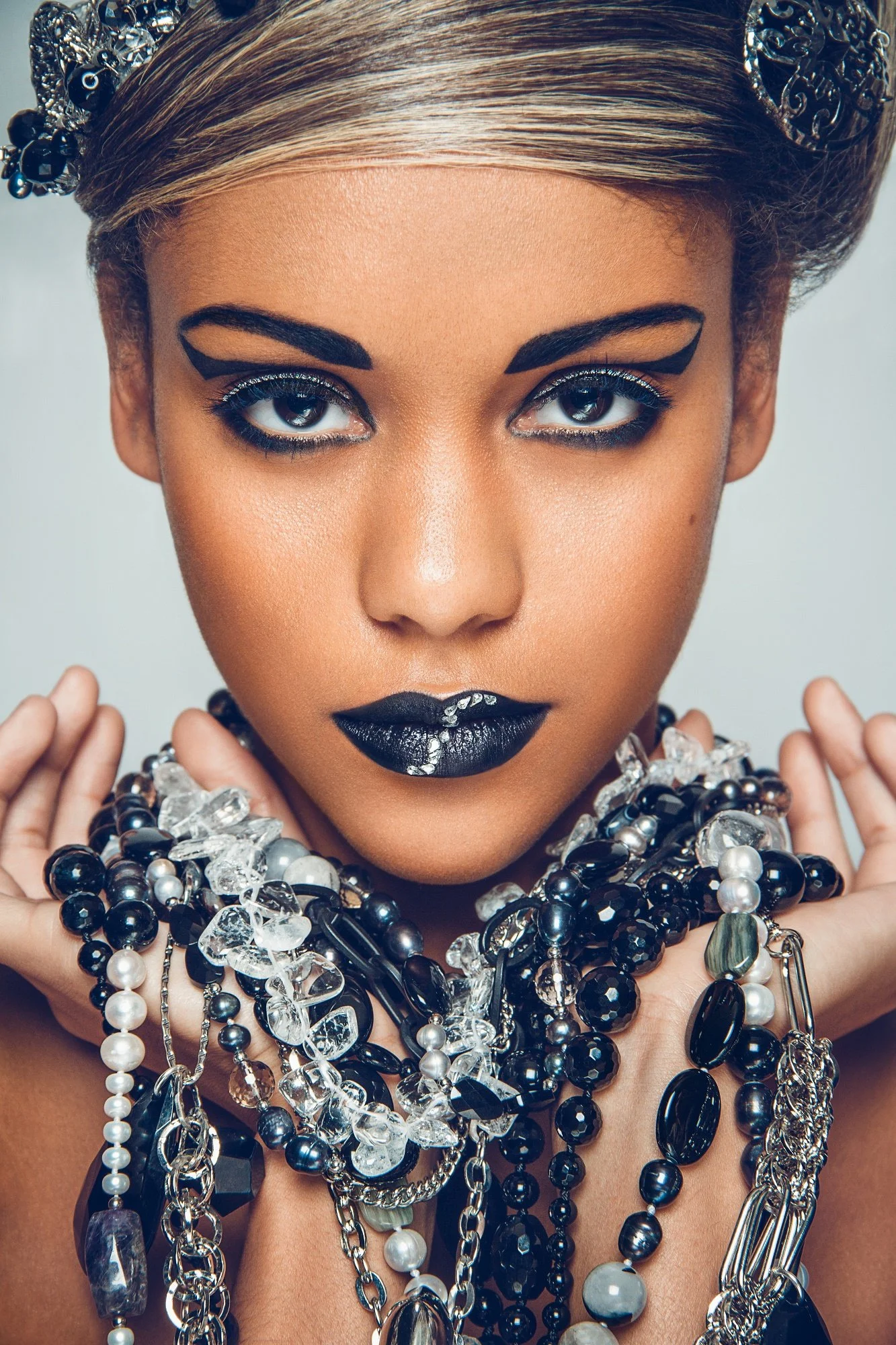 A woman with bold black and silver makeup, dark lipstick, and wearing numerous black, clear, and pearl jewelry necklaces.