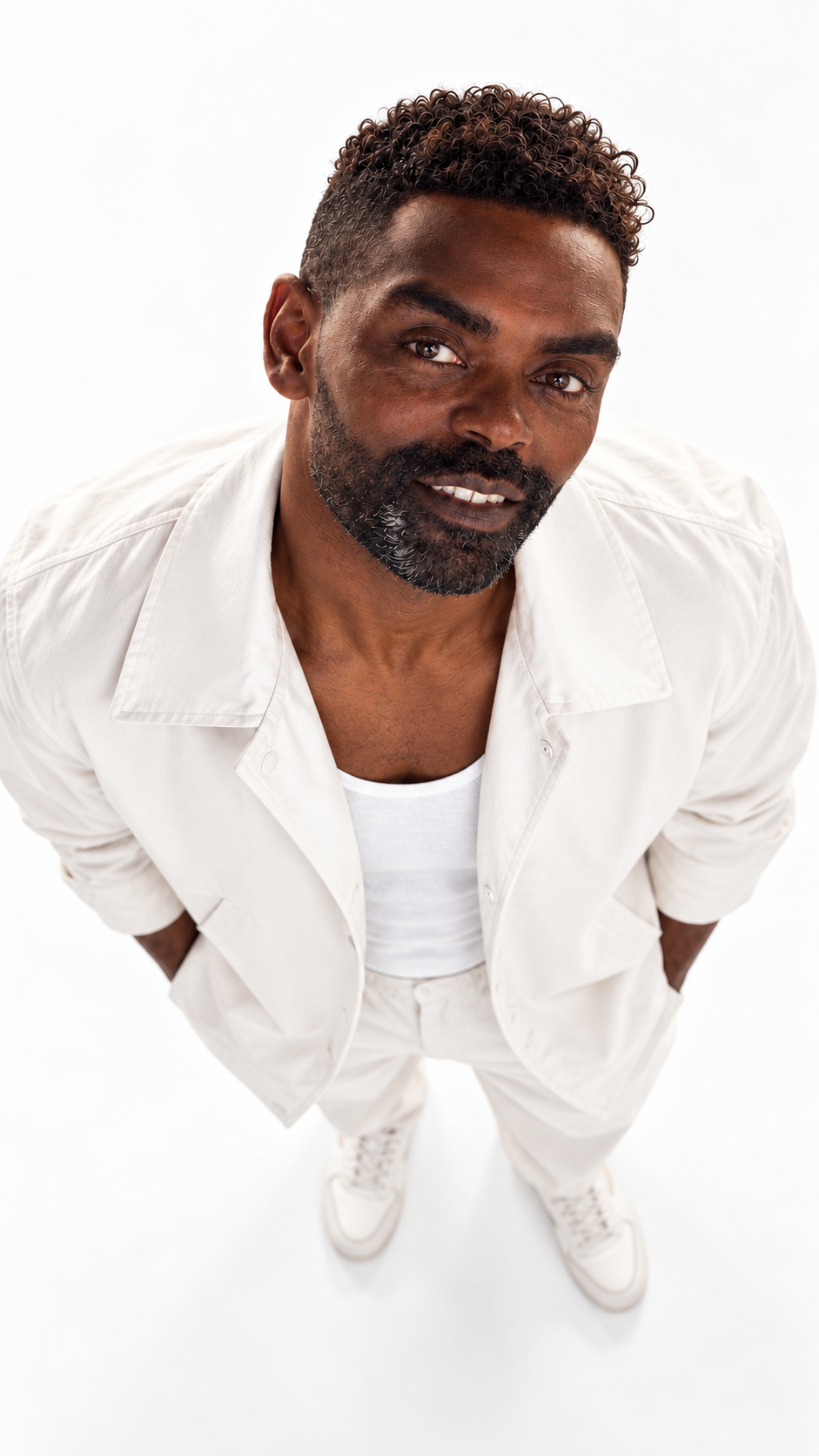 A man with short, curly hair and a beard, dressed in a white shirt, white t-shirt, and white sneakers, standing against a white background and looking at the camera.