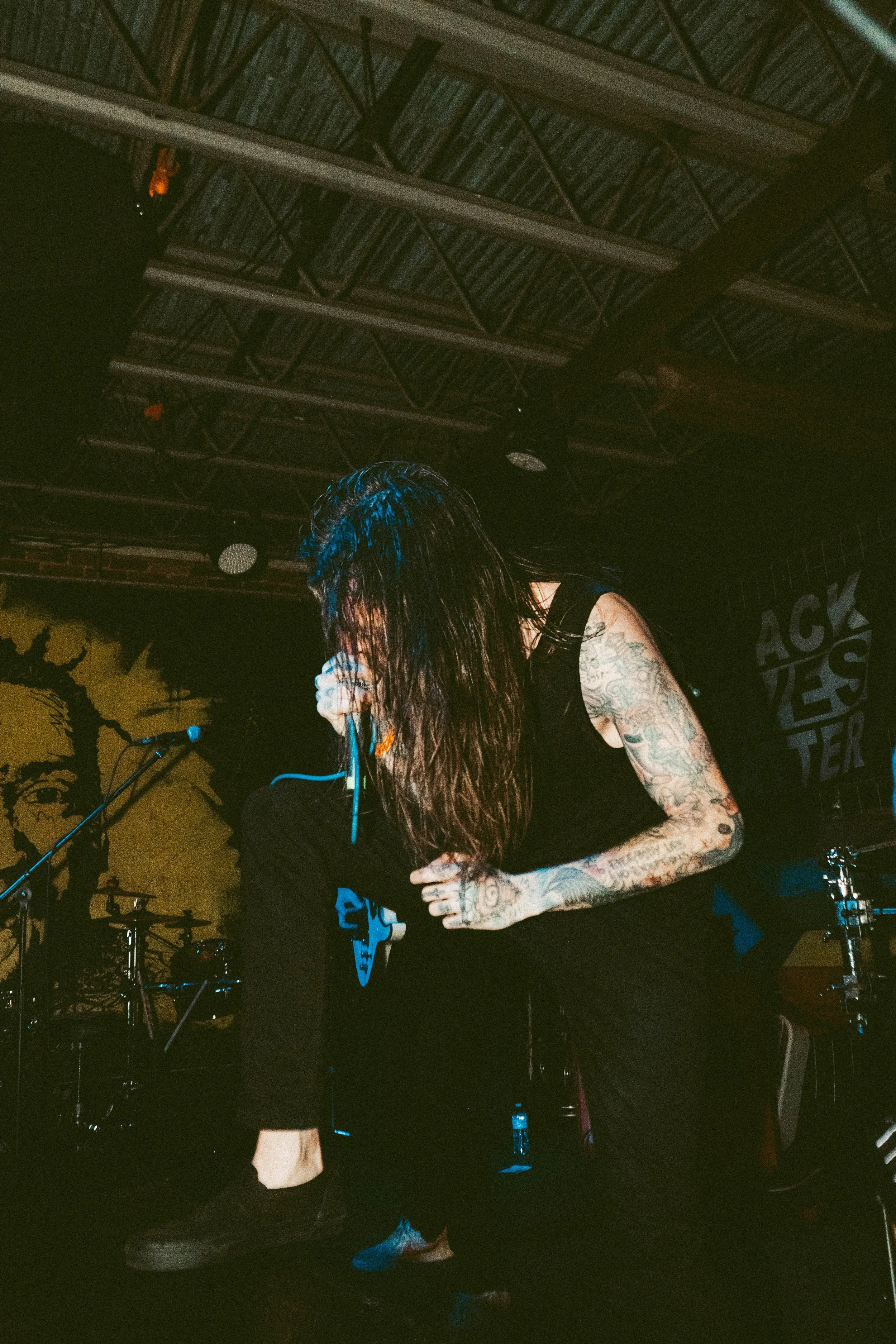 Male singer with long dark hair and tattoos on her arms, singing into a microphone on a dimly lit stage with musical equipment and drum set in the background.