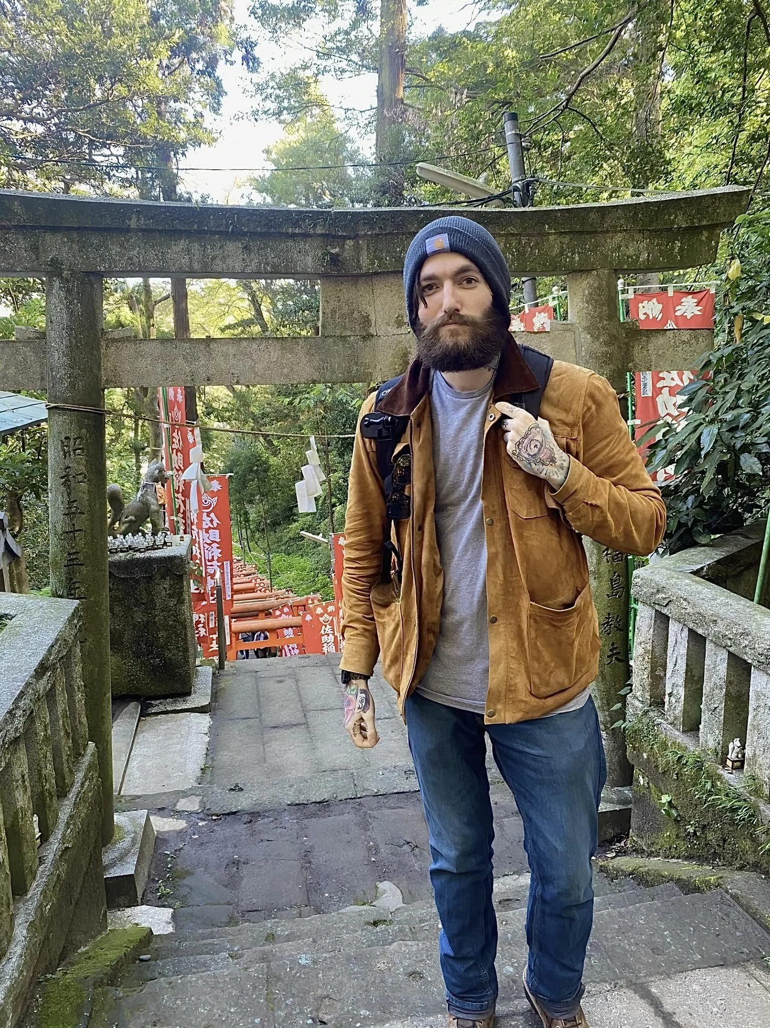 A man with a beard, tattoos, wearing a blue beanie, tan jacket, gray shirt, blue jeans, and carrying a backpack, standing on stone steps in front of a traditional Japanese torii gate with moss and greenery around.