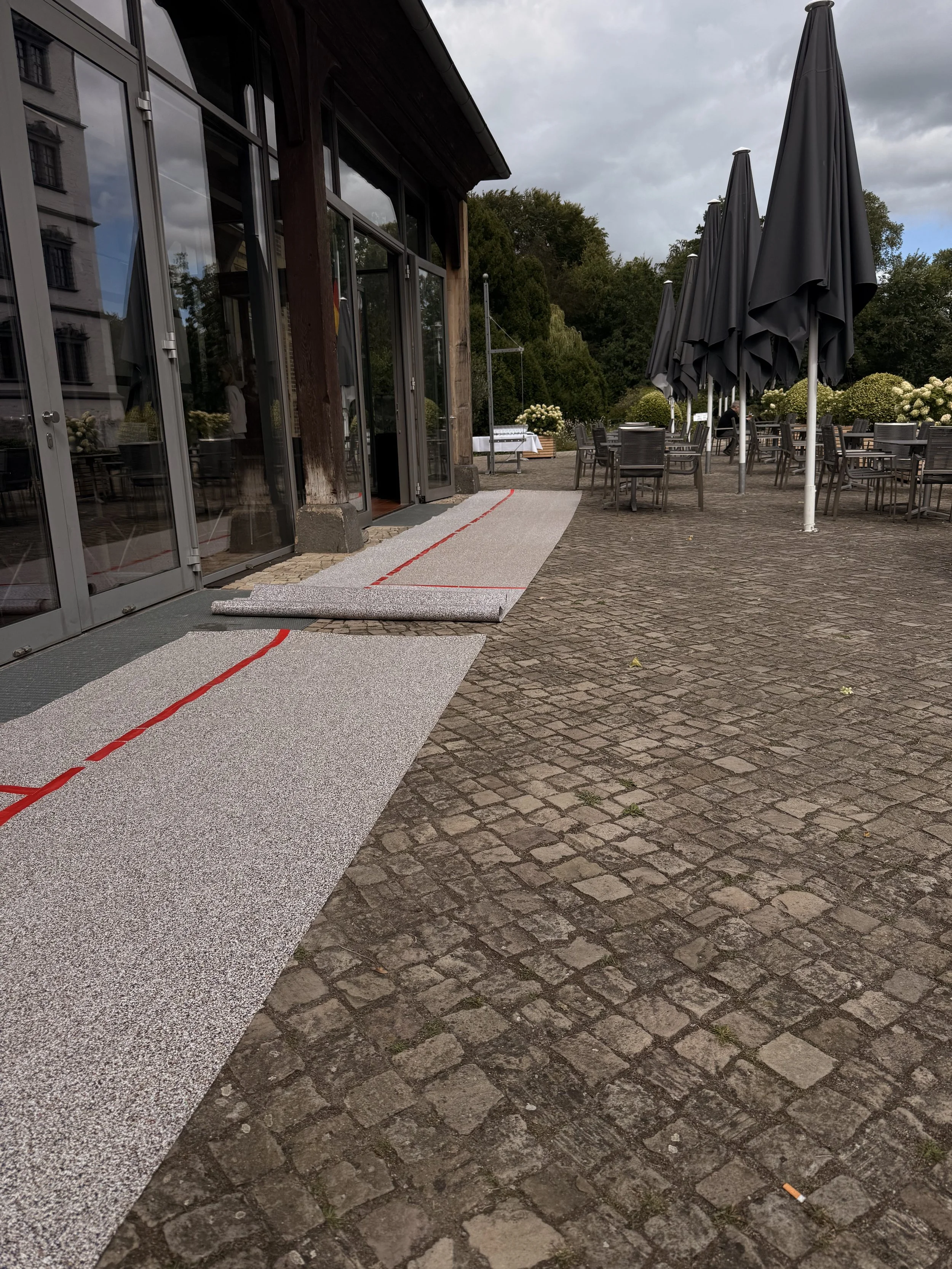 Die Terrasse vor einem Gebäude mit gläserner Tür und mehreren schwarzen Sonnenschirmen und Tischen. Der Boden besteht aus Pflastersteinen, einige Blätter und eine Zigarette liegen herum.