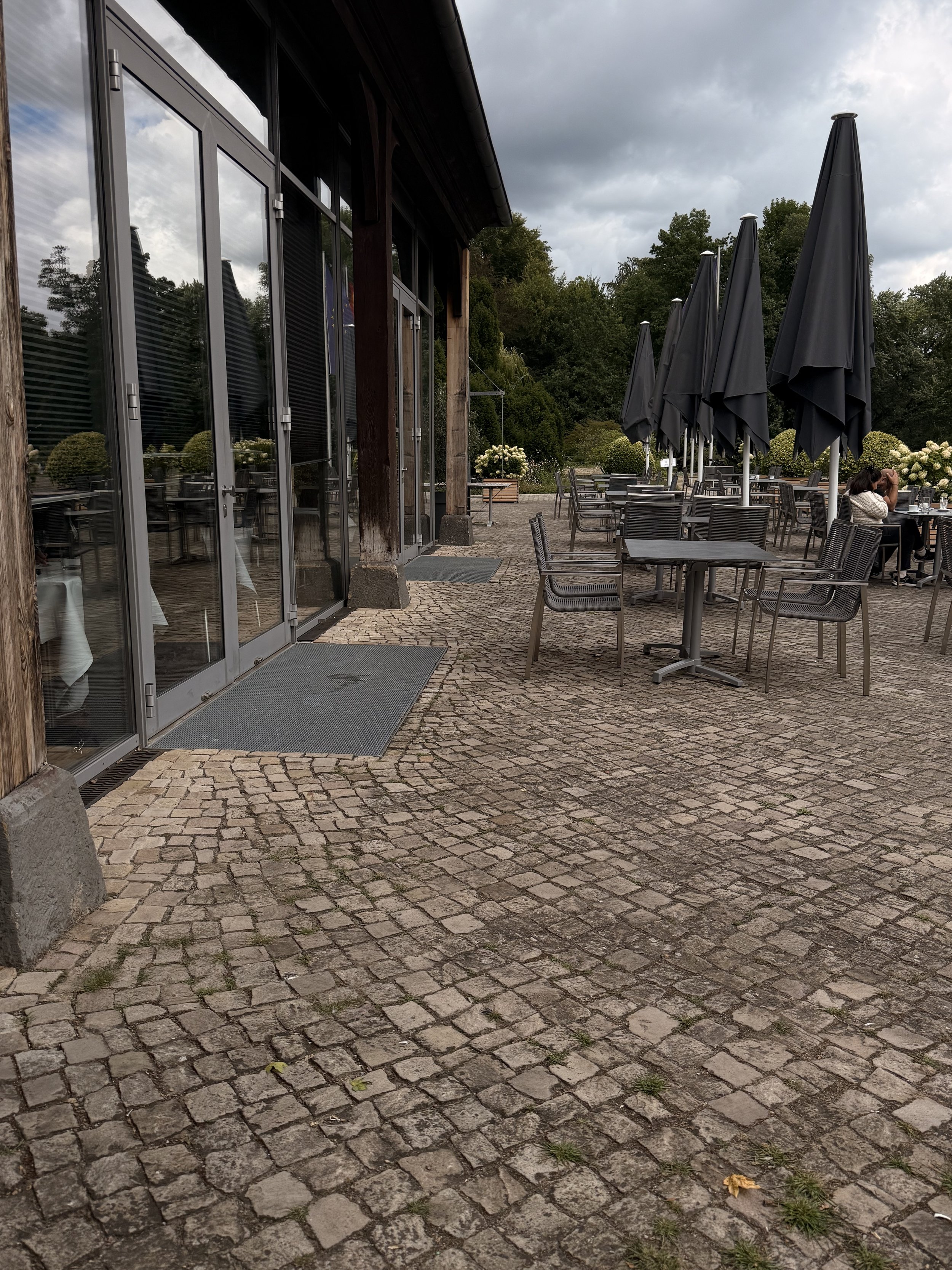 Außenaufnahme eines Restaurants im Freien mit mehreren schwarzen Sonnenschirmen, Tischen und Stühlen, und einem Paar, das sich unter einem Schirm küsst. Der Boden ist gepflastert, im Hintergrund sind Bäume und ein bewölkter Himmel sichtbar.
