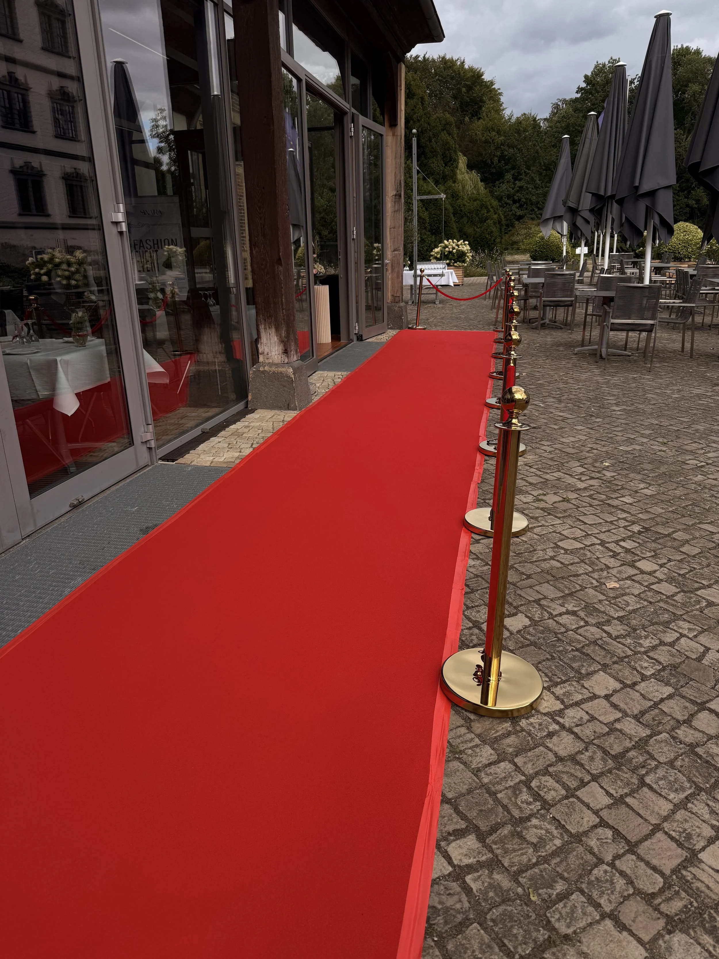 Verkaufslale mit rotem Teppich, rotes Seil und goldene Pfosten, im Freien sitzende Tische und Stühle unter dunklen Schirm, sonnige und bewölkte Himmel, Bäume im Hintergrund.