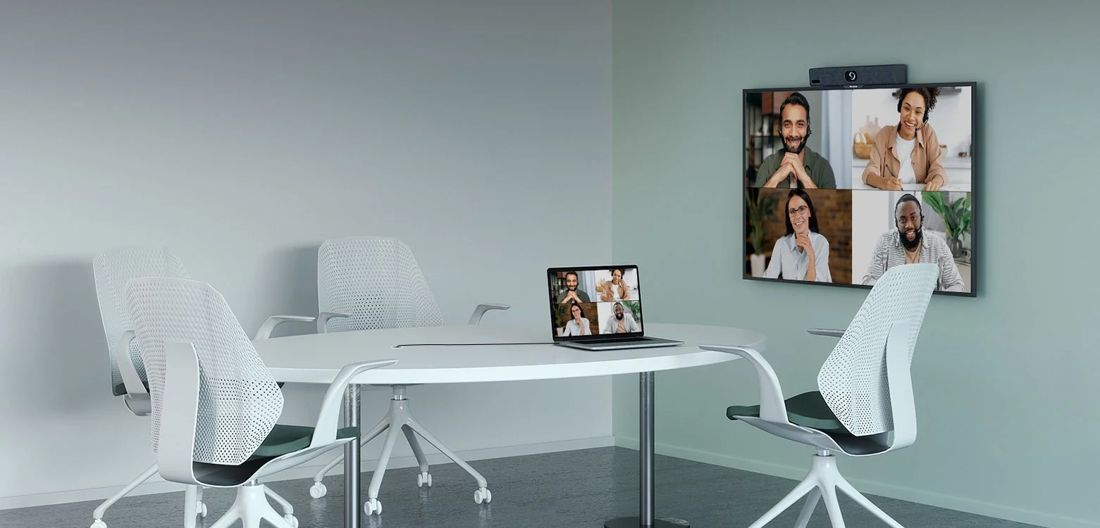 Virtual meeting room with a white oval table, four white mesh office chairs, a laptop displaying a video call, and a mounted screen showing the same video call with five participants, in a modern office setting.