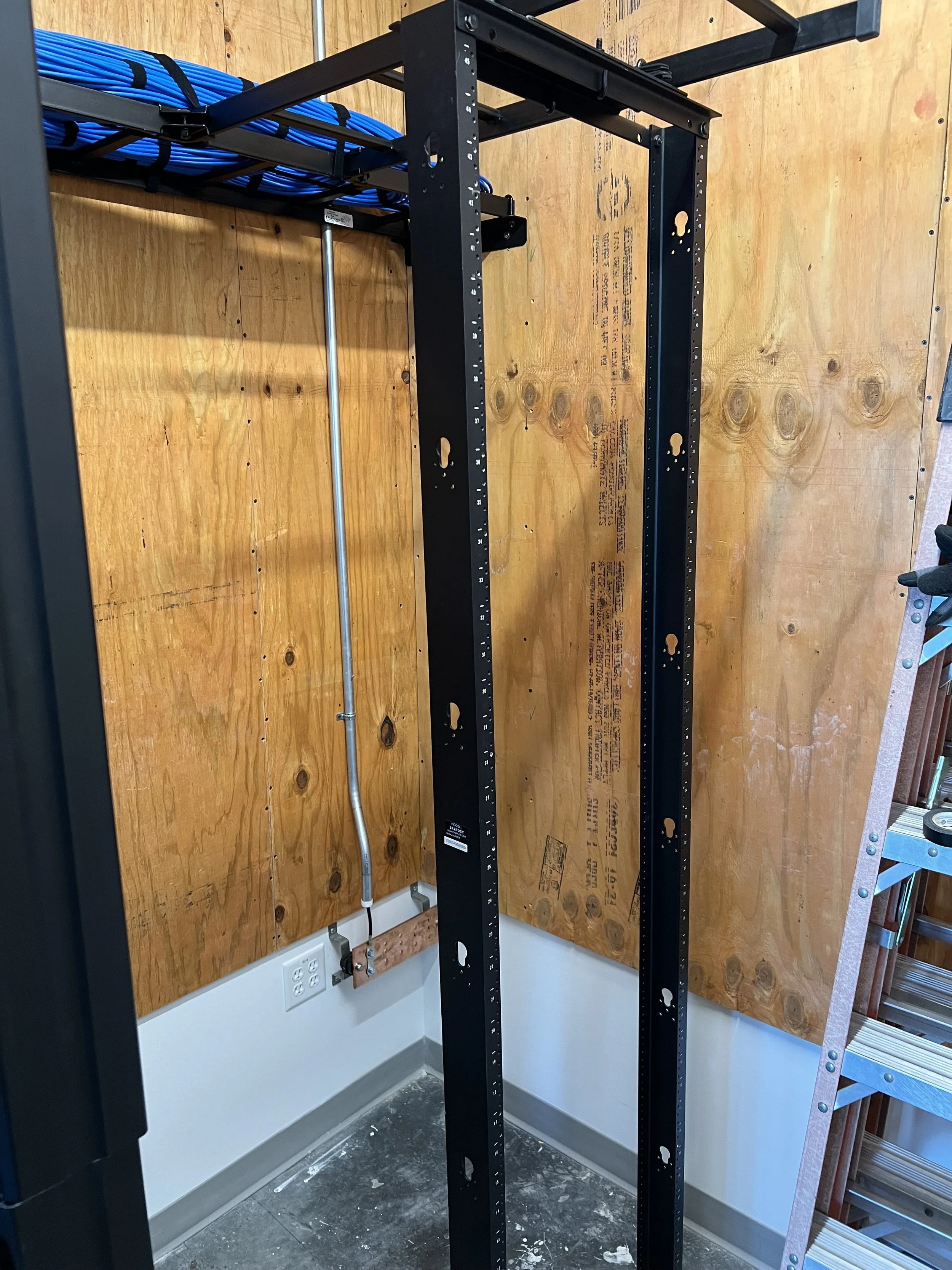 Empty black metal shelving unit in front of a wooden wall with a bundle of blue cables secured on a rack and a power outlet on the wall.