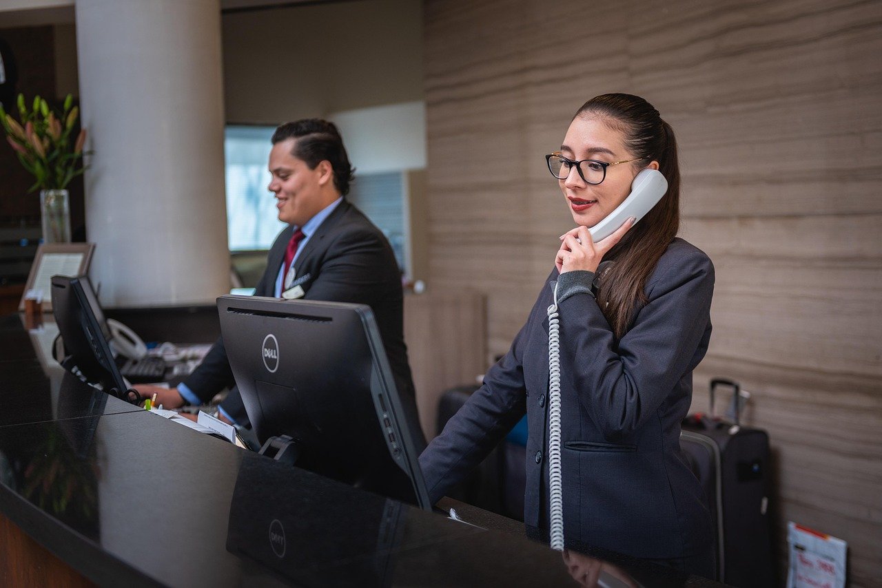 Hotel reception desk with a female receptionist talking on the phone and a male receptionist working on the computer, in a modern hotel lobby.
