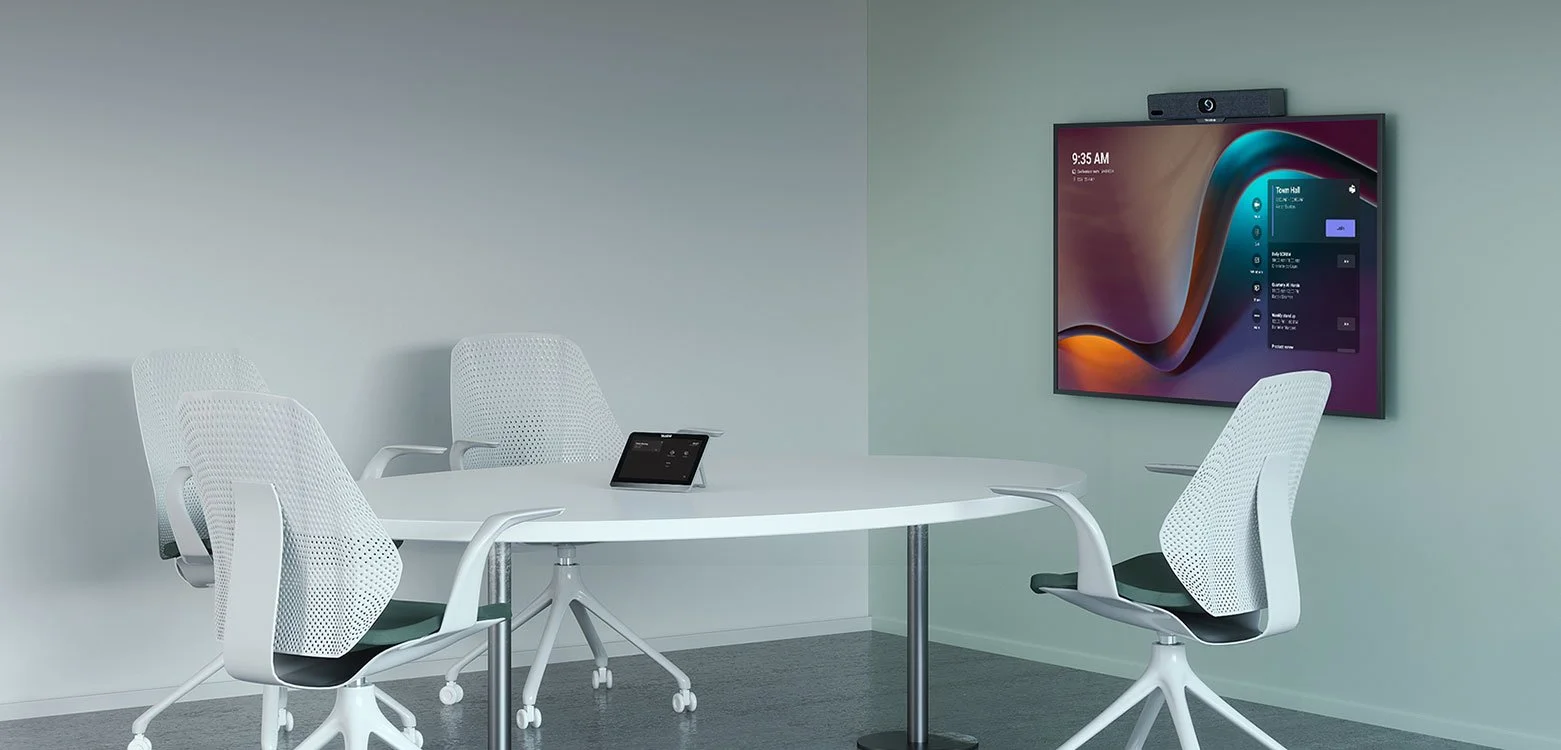 A modern conference room with three white office chairs around a white oval table, a tablet on the table, and a large wall-mounted screen displaying a colorful graphical interface, with a video conferencing camera above the screen.