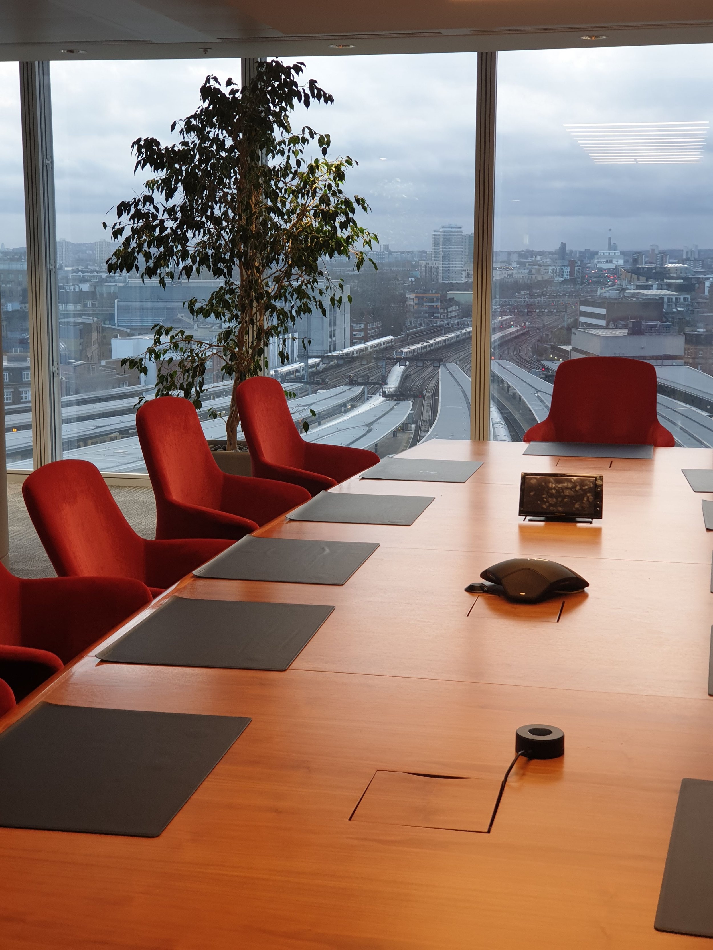 Conference room with a large wooden table, red chairs, a tablet, a computer mouse, a travel mug, and a large window showing cityscape and train tracks outside.