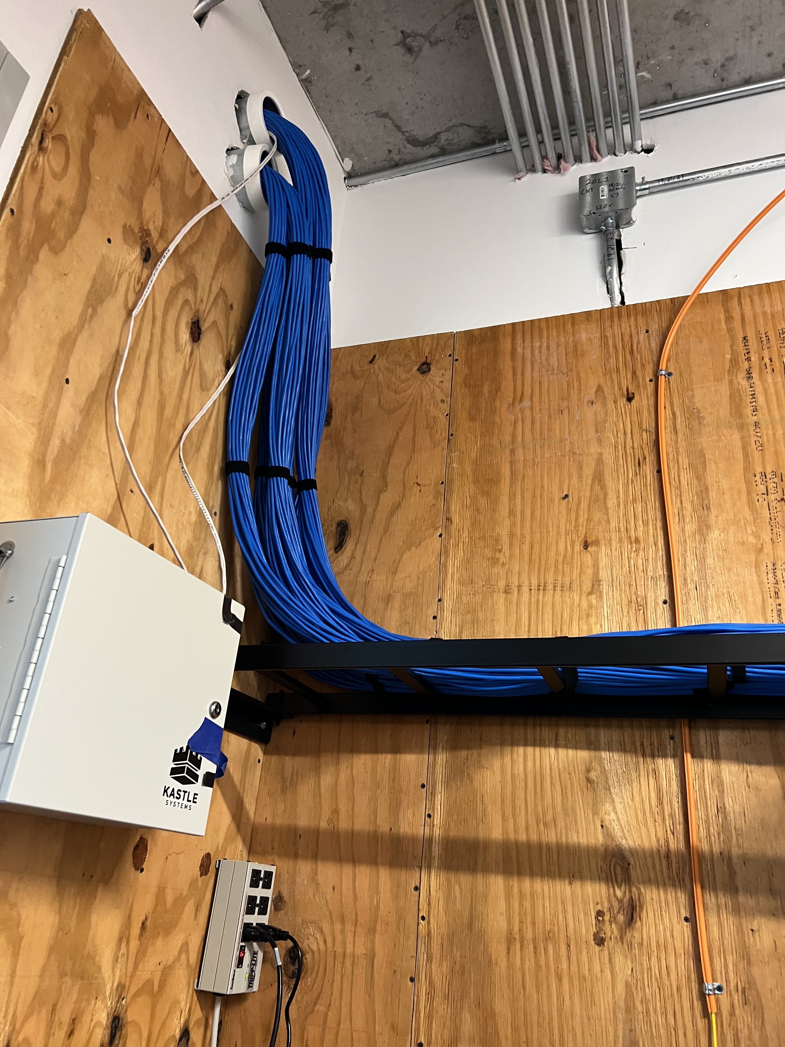 Electrical wiring and network cables organized behind a wall, with a power strip, electrical outlets, and a box labeled "Kastle Systems."
