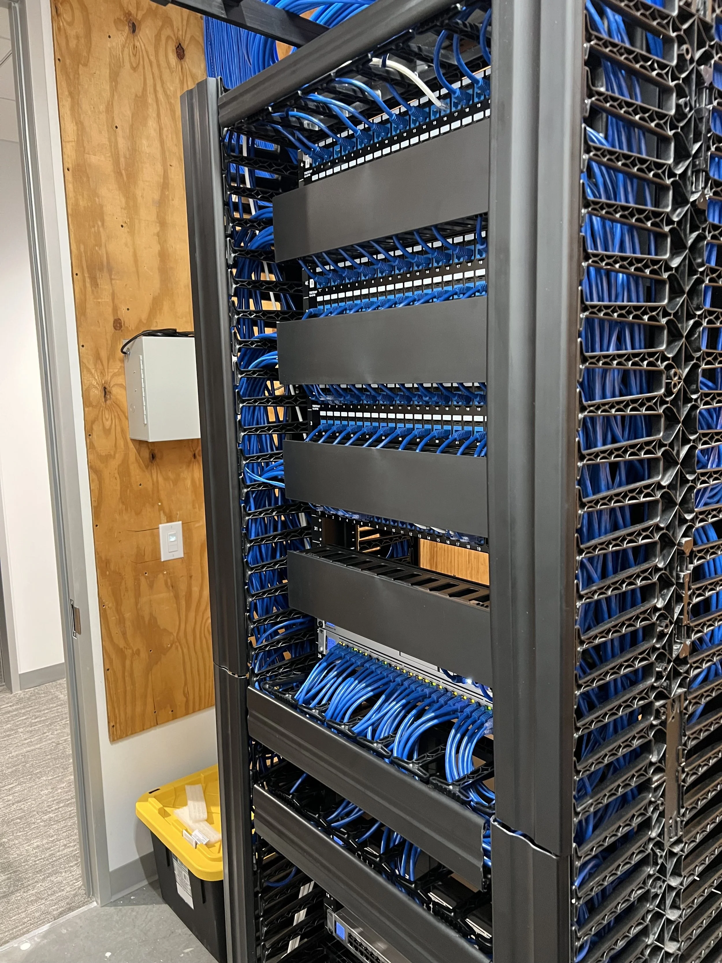 A server rack filled with blue Ethernet cables in a data center, with a white wall and wooden panel in the background.