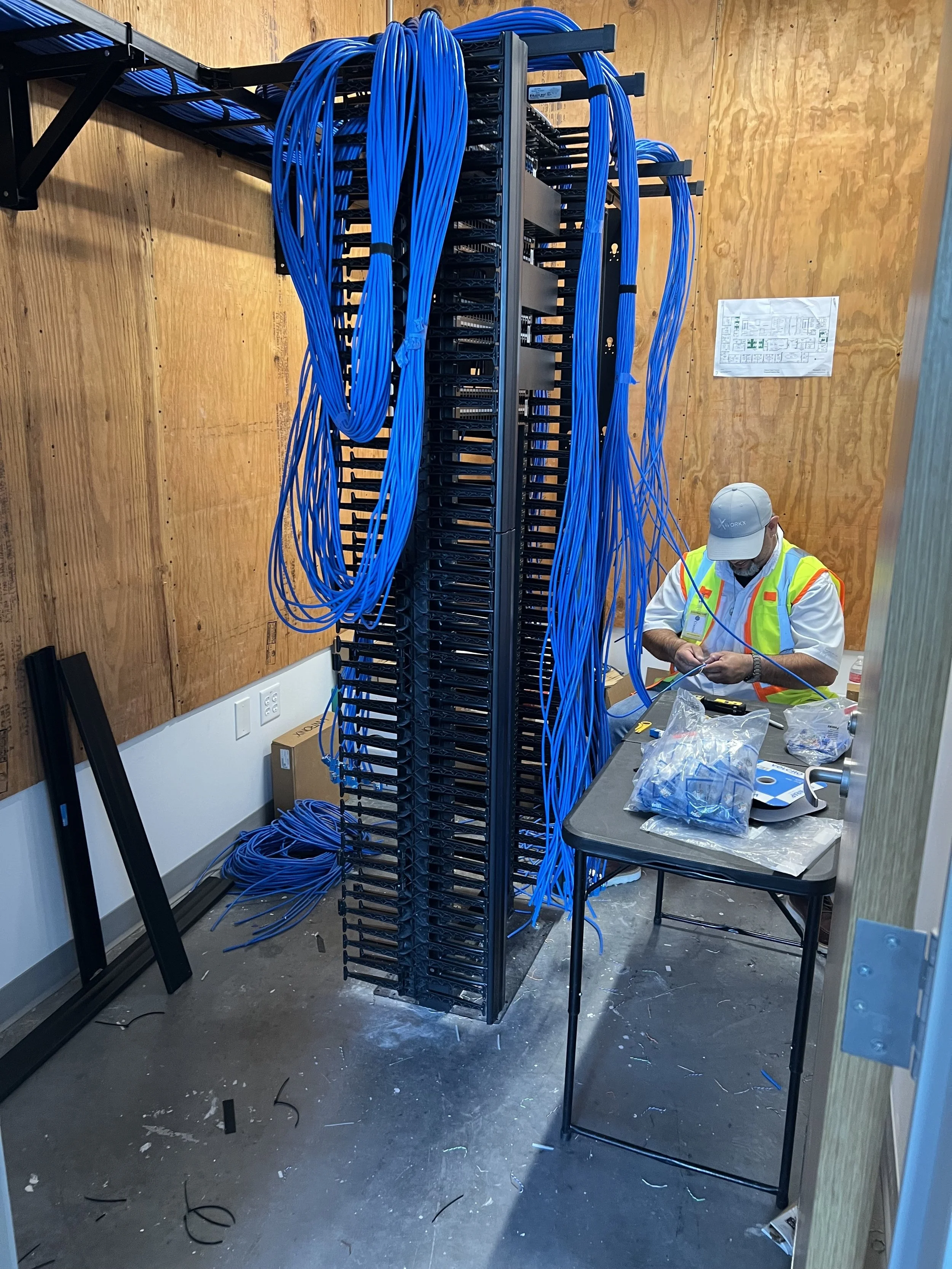 Technician working on a large server rack with blue Ethernet cables, in a room with wooden walls and a table with equipment and tools.