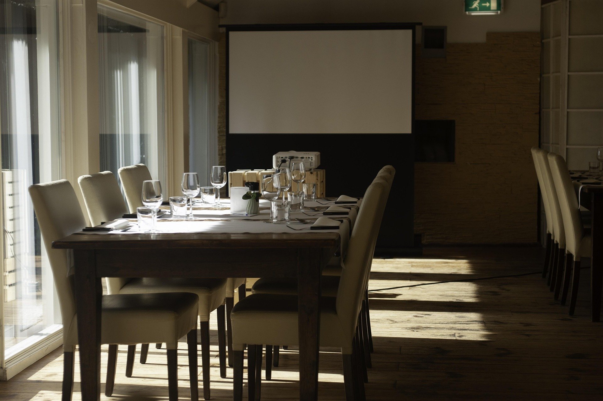 A dining area with a long table set with wine glasses, plates, and cutlery, surrounded by cream-colored chairs, and a projector screen in the background. Sunlight streams through sliding glass doors on the left.