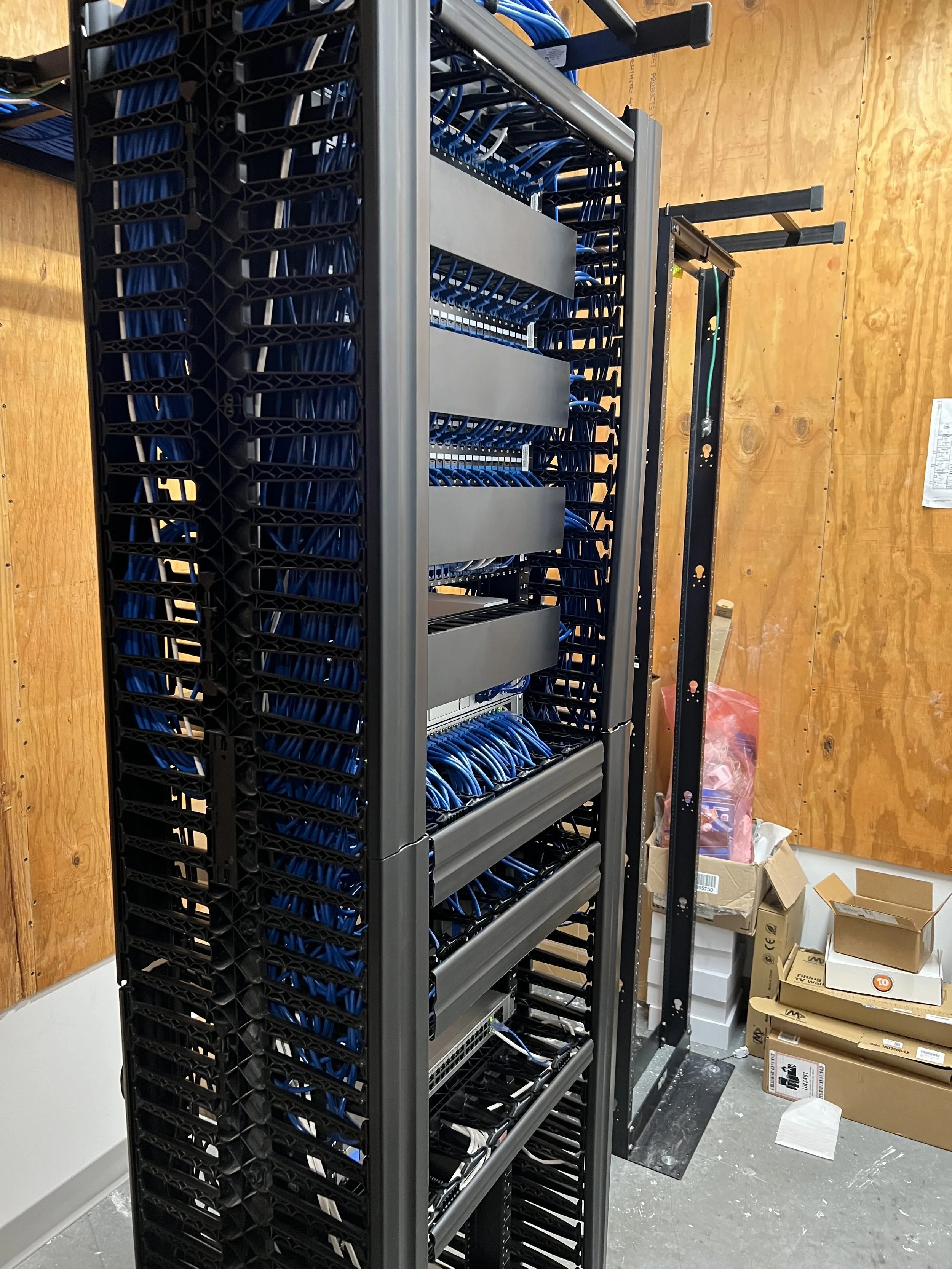 A server rack with multiple network switches and blue Ethernet cables in a data center or server room, with some boxes and packaging materials on the floor nearby.