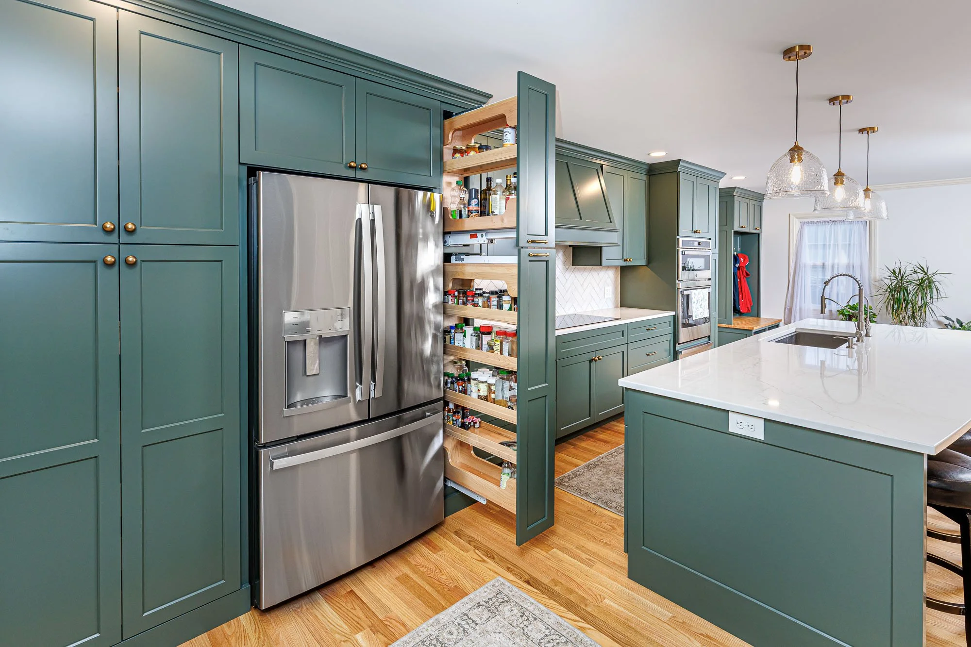 Modern green kitchen cabinets with a stainless steel refrigerator and open spice cabinet, white island with a built-in sink, hardwood flooring, and hanging light fixtures.
